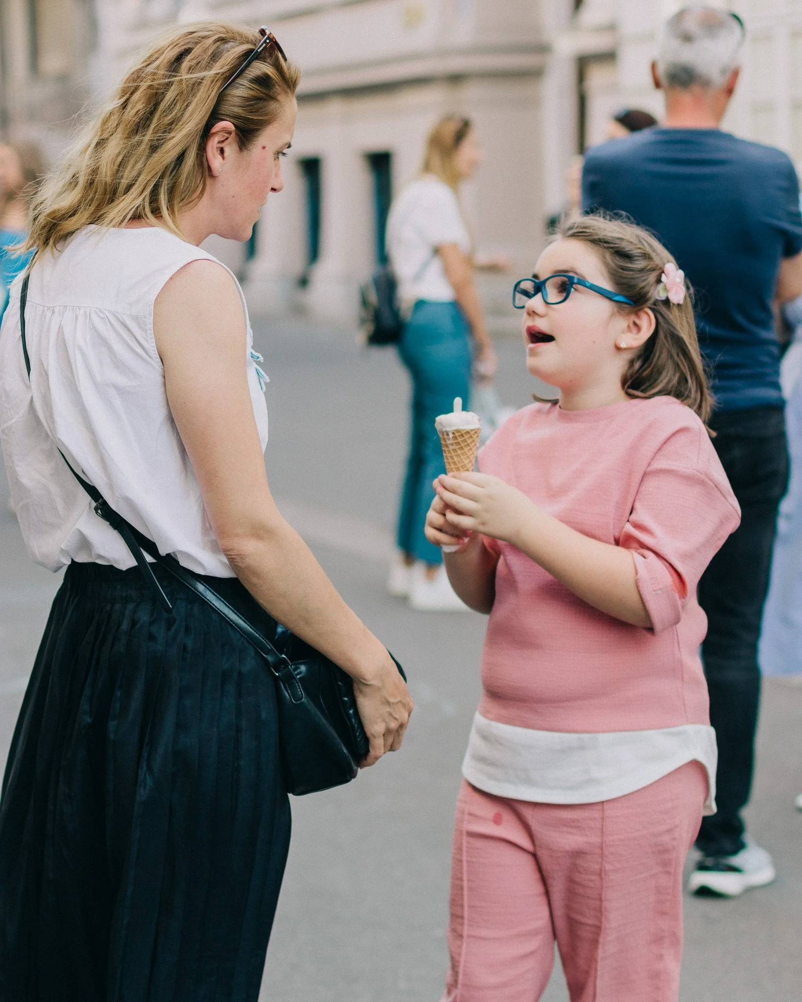 A woman and a girl with glasses are standing on a street, having a conversation. The girl is holding an ice cream cone and wearing a pink outfit, while the woman is dressed in a white top and black skirt.