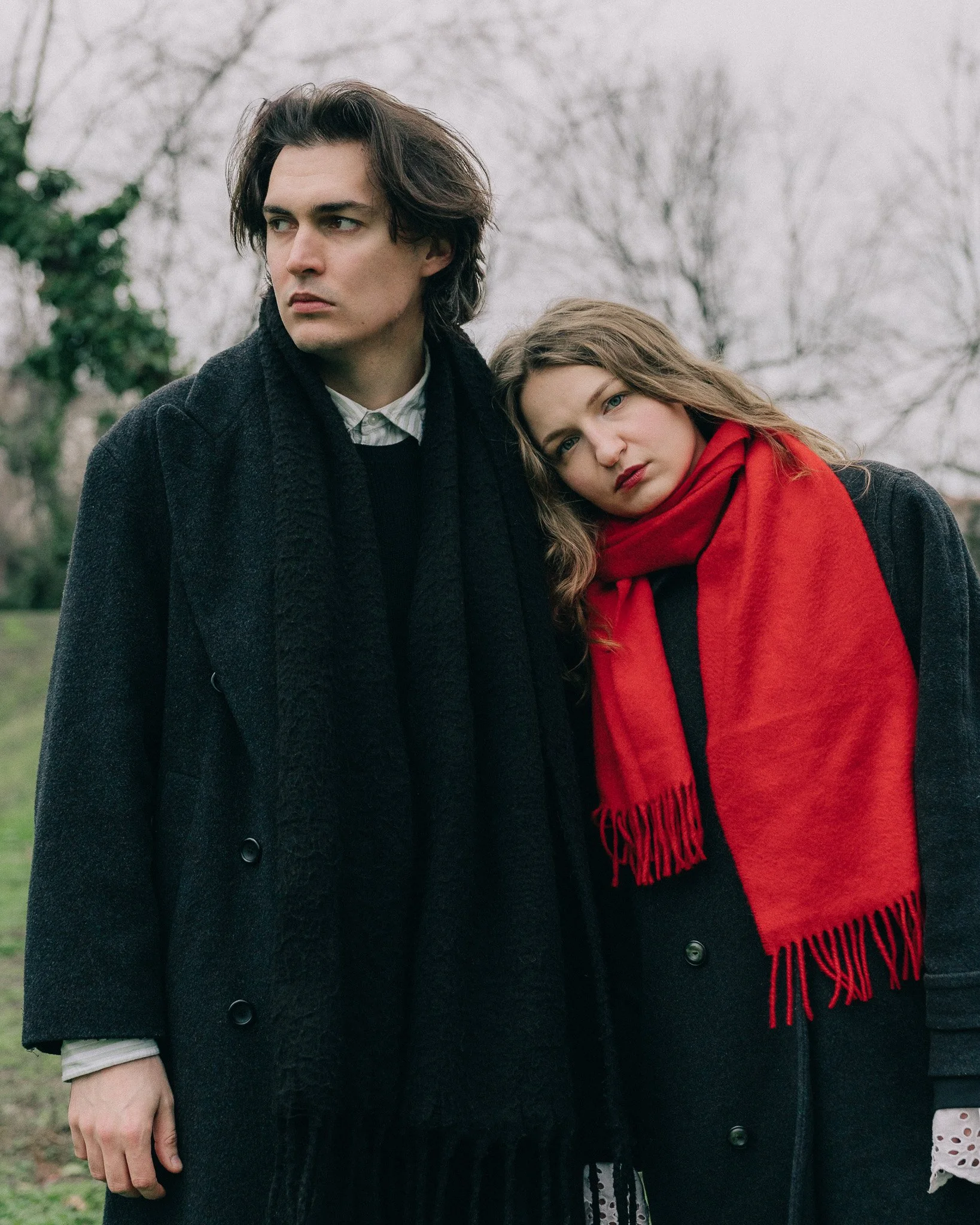 A young man with dark brown, wavy hair and a young woman with long, wavy blonde hair leaning against each other outdoors on a cloudy day, both wearing dark coats, with the woman also wearing a bright red scarf.