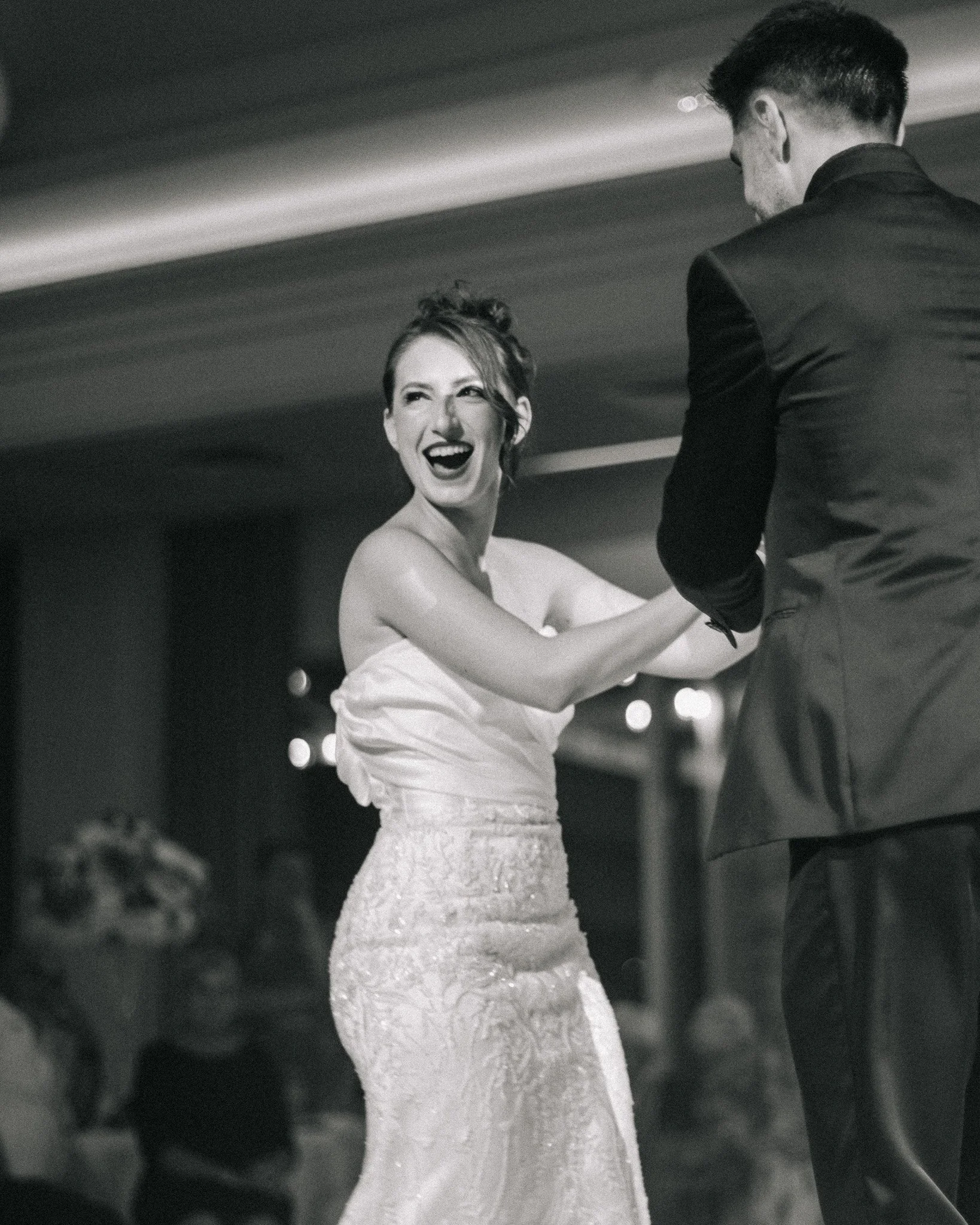 A woman in a wedding dress laughing and dancing with a man in a tuxedo at a celebration.