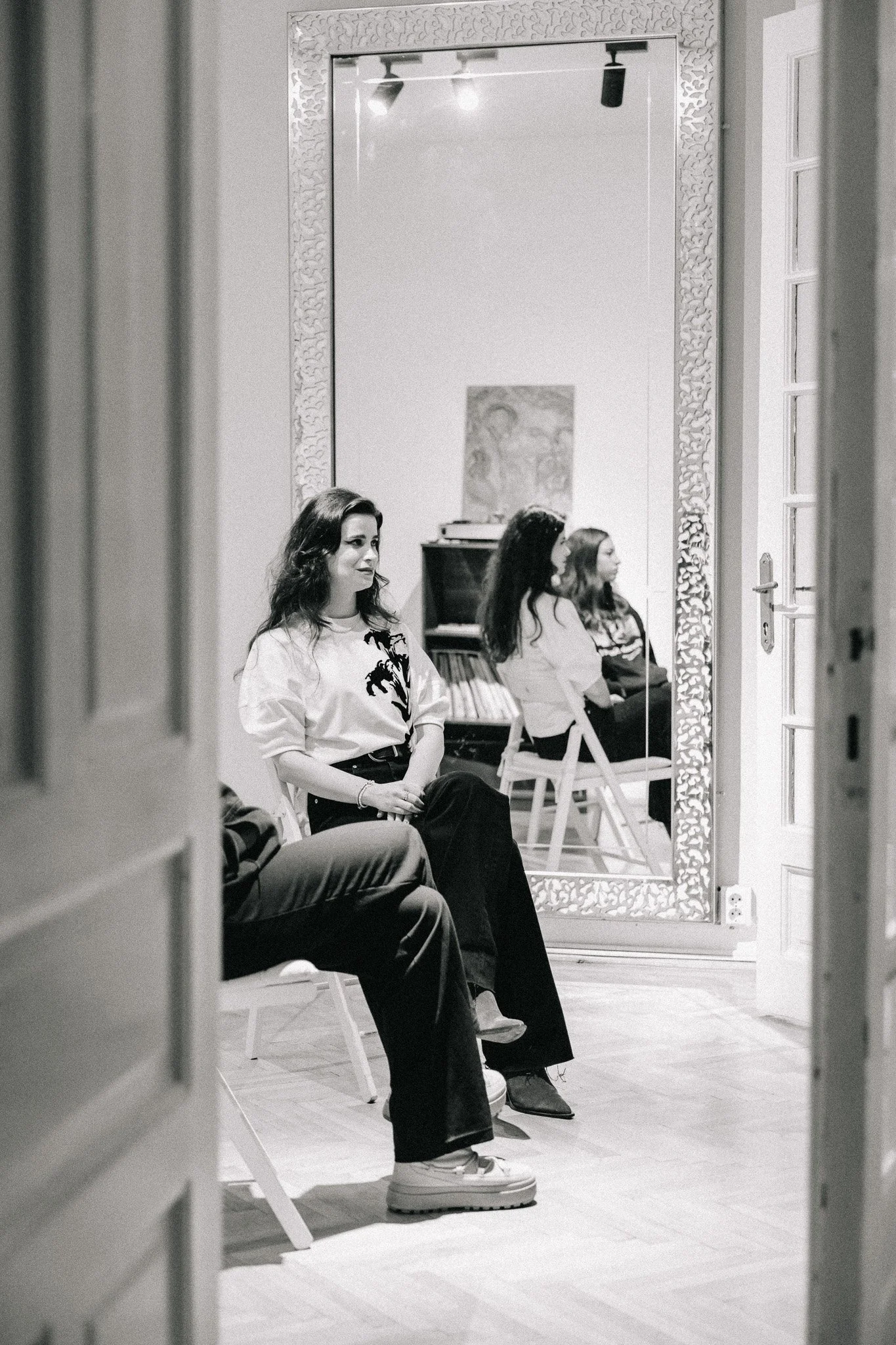 A woman sitting on a chair in a room, reflected in a large mirror with ornate border; two other women are seated in the background, one facing the mirror and the other with her side to the camera.