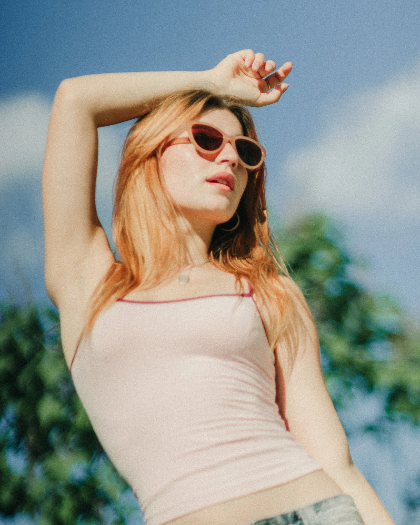 A woman with red hair wearing sunglasses and a beige tank top outdoors with trees and a blue sky in the background.