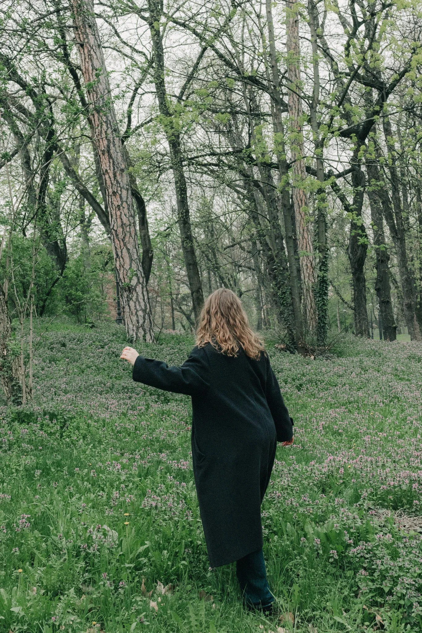 A woman with curly hair wearing a long black coat walking through a lush green meadow with small pink and white flowers, surrounded by tall trees in a forest.