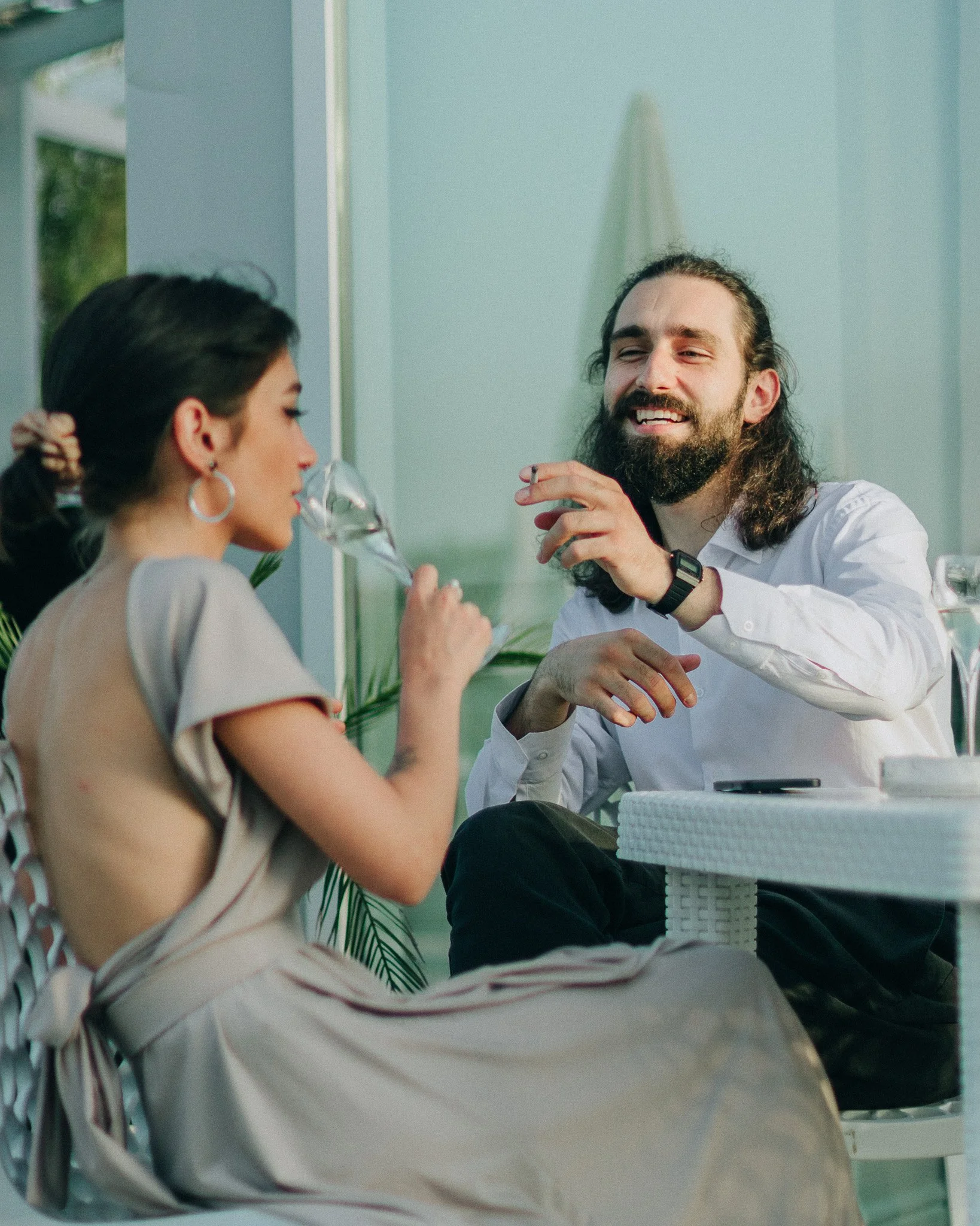 A man with long hair and a beard smiling while talking to a woman drinking from a glass of wine in an outdoor setting.