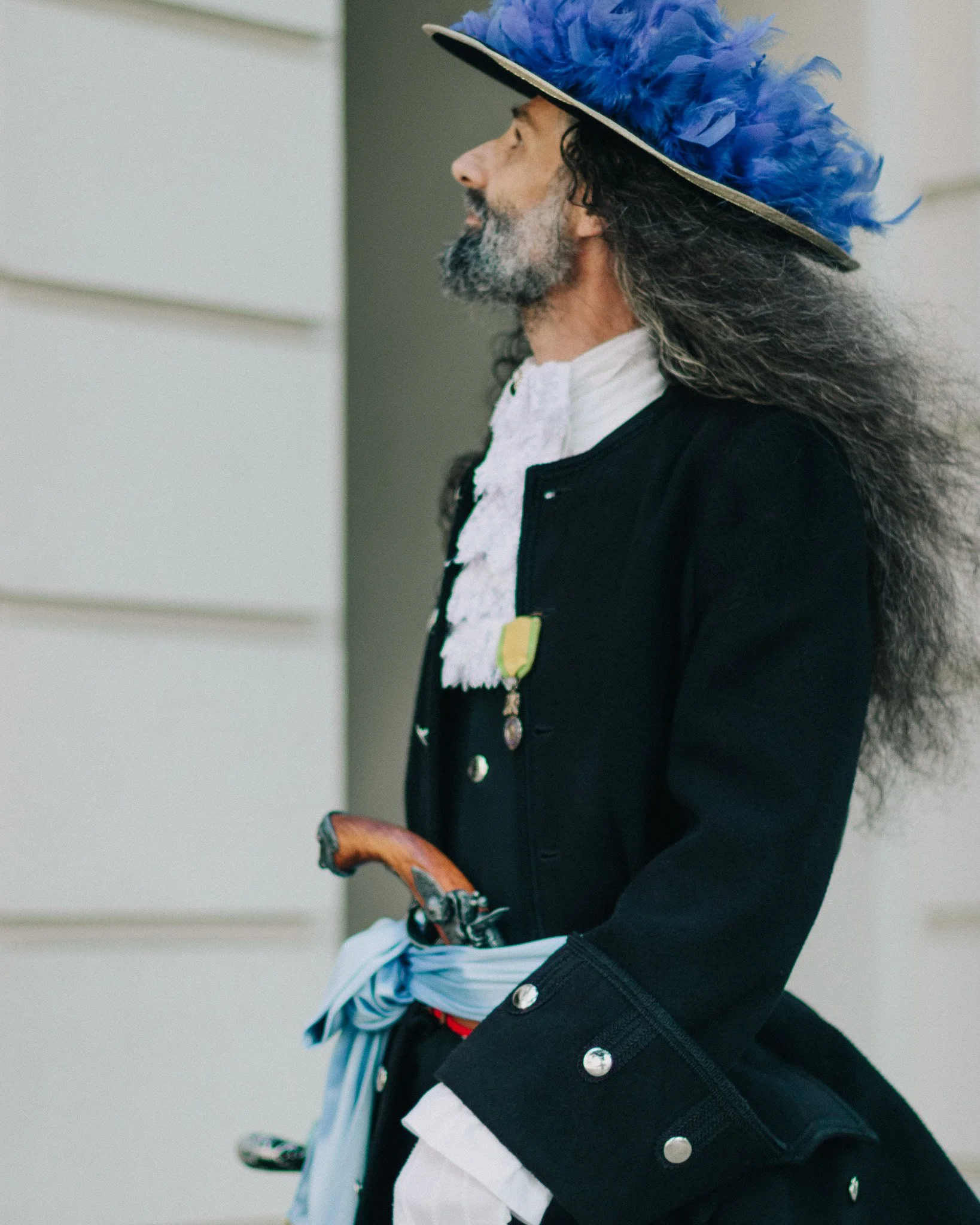 A man with long gray hair and a gray beard wearing a colorful hat with blue feathers, vintage clothes, and holding a cane, standing outdoors.