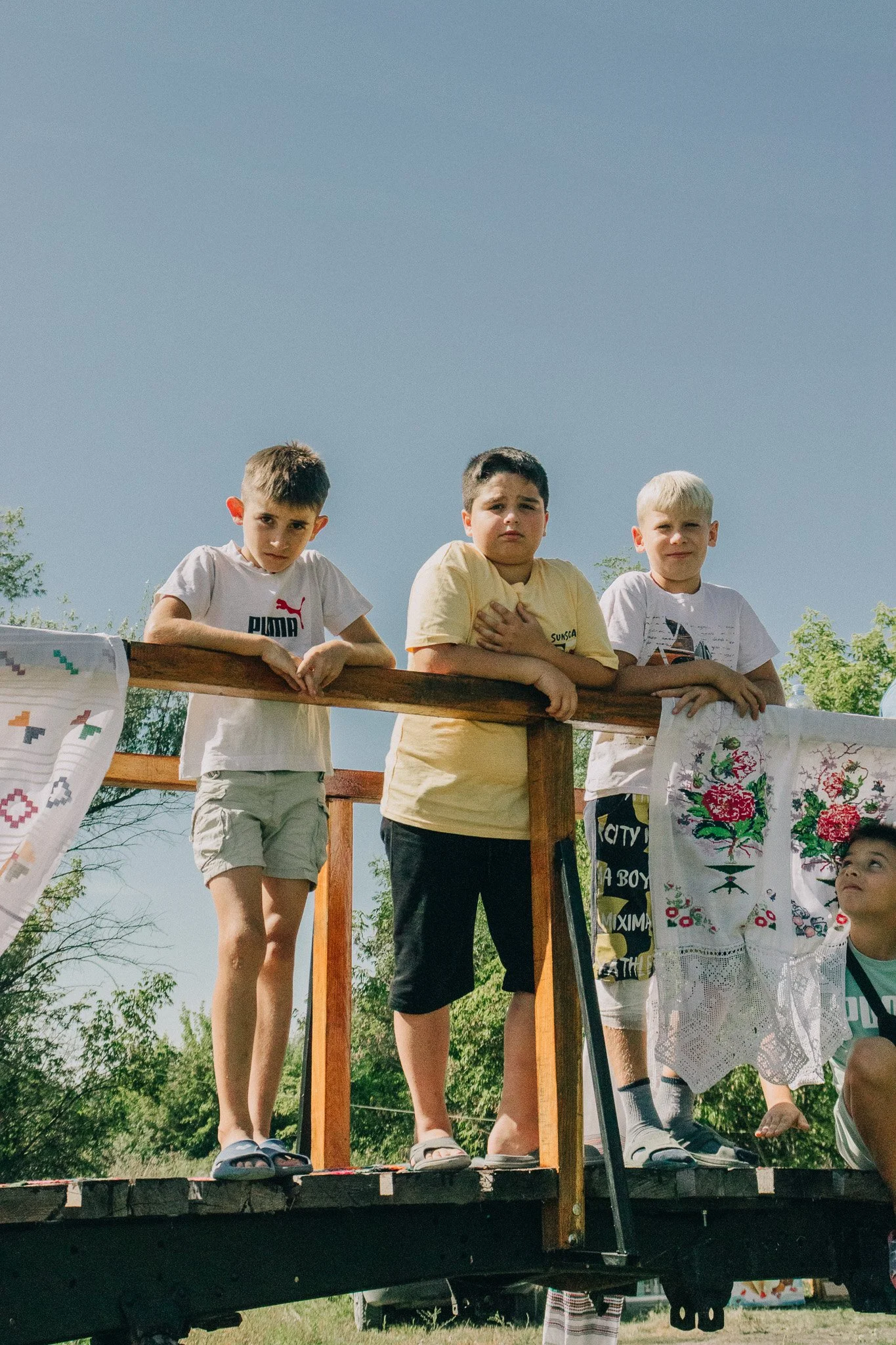 Group of children standing on a wooden balcony outdoors, some with fabric embroidered textiles hanging on the railing, against a clear blue sky.