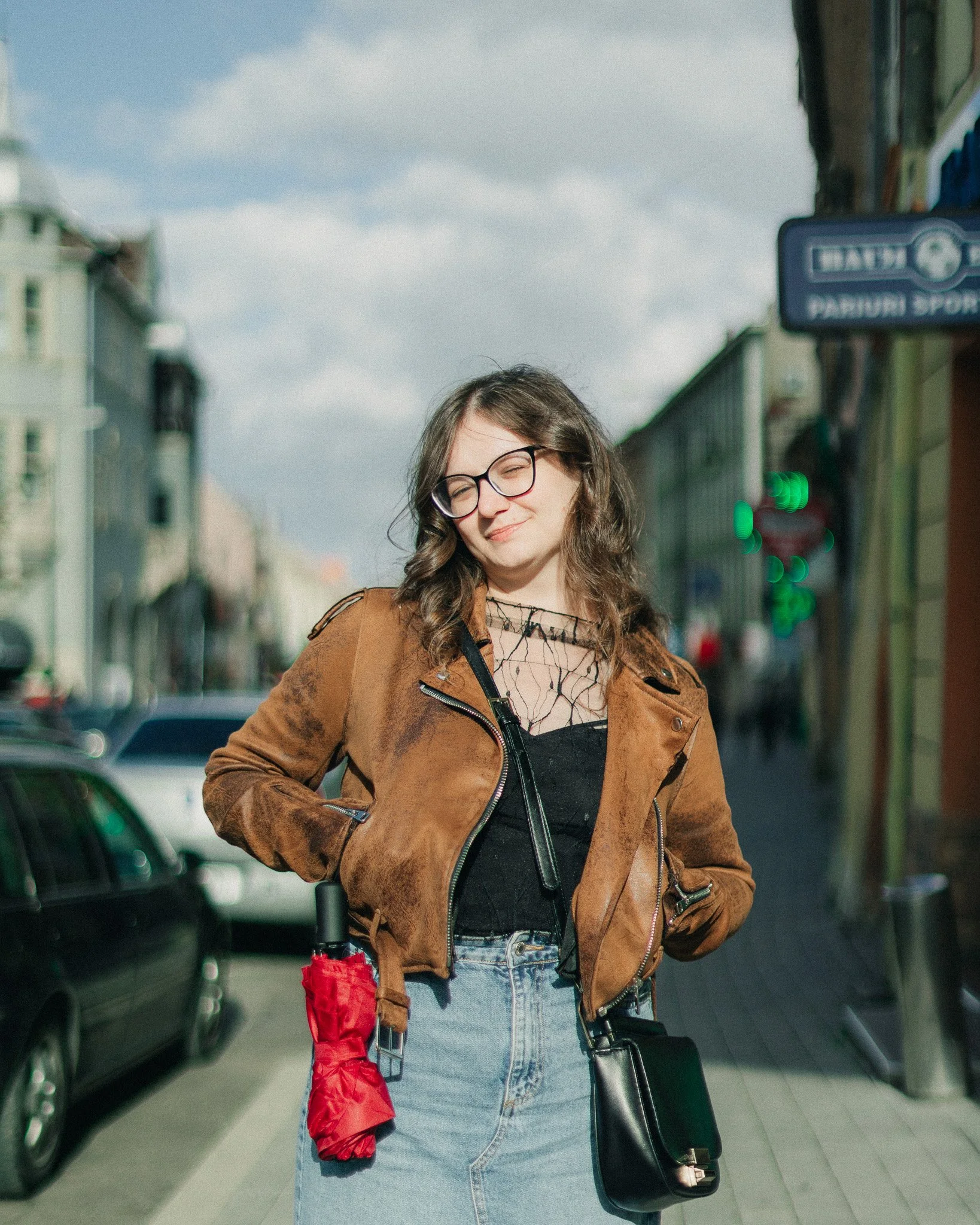 Young woman with glasses smiling on city street, holding red umbrella, wearing brown leather jacket and light blue jeans.