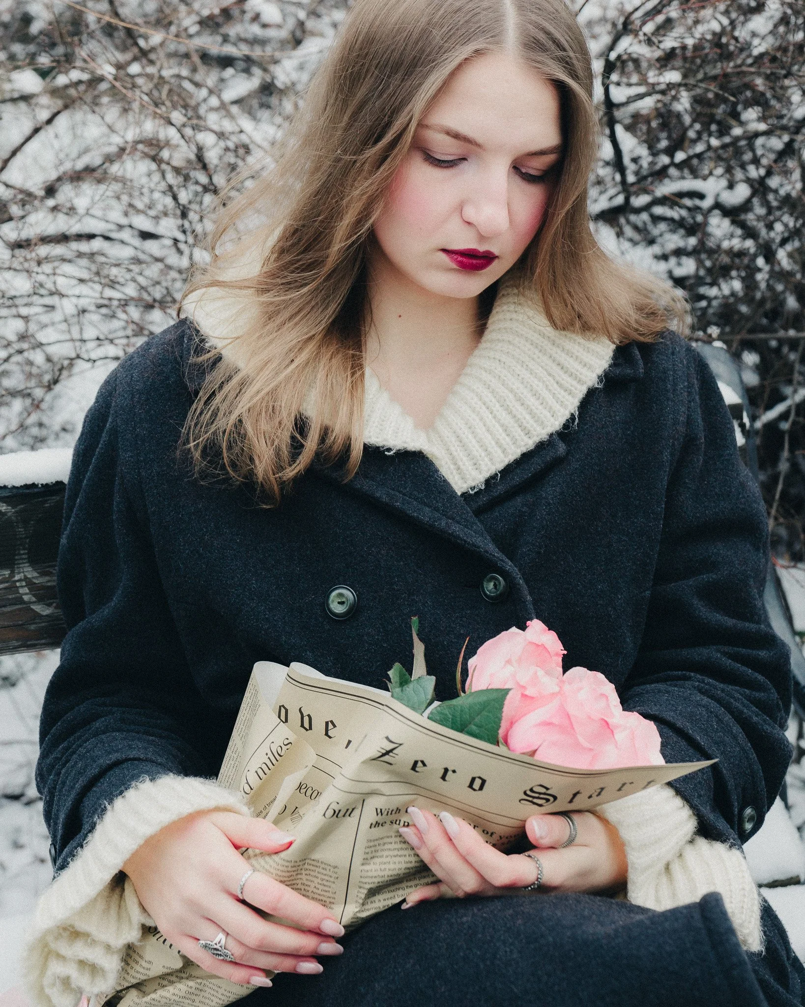 A young woman with light brown hair, wearing a dark coat and a cream-colored sweater, is sitting outdoors in a snowy setting, reading a newspaper and holding a pink rose.