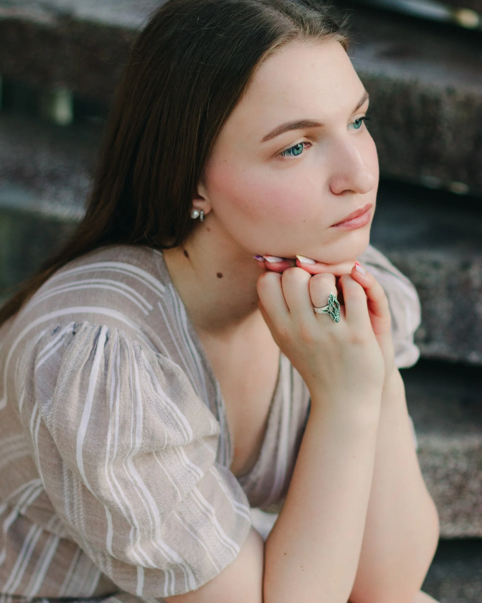 A young woman with long brown hair, blue eyes, and fair skin, resting her chin on her hands, sitting outdoors near a wooden structure, wearing a striped beige and white top, and jewelry including earrings and a large ring.