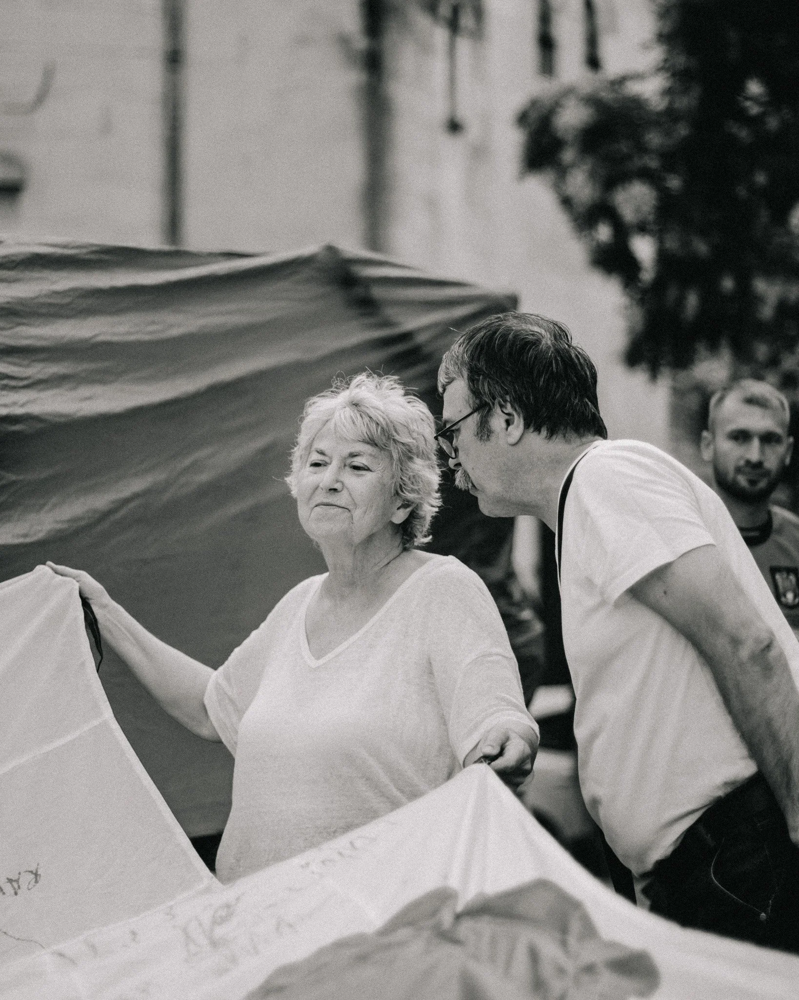A black and white photo of an elderly woman, a younger man, and a man in the background, all holding umbrellas on a street. The elderly woman has short, wavy hair and is wearing a light-colored top, while the younger man has dark hair, glasses, and i