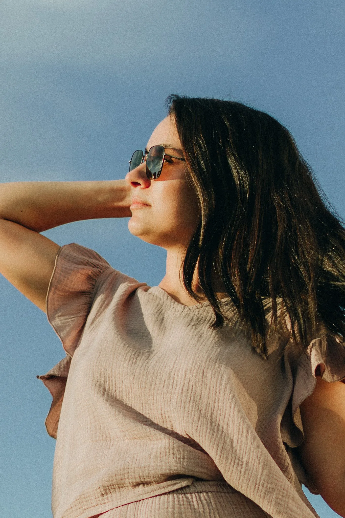 A woman with dark hair wearing sunglasses and a beige top, standing outdoors against a blue sky with sunlight on her face.
