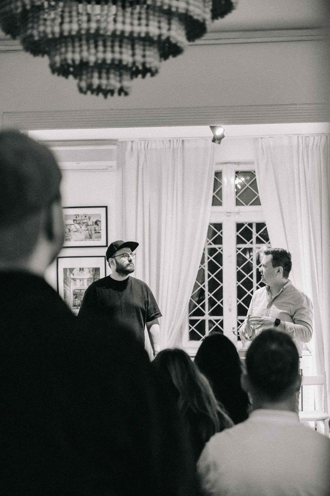A black and white photo of a group gathering in a room with two men standing at the front, speaking or presenting to an audience. The room has tall windows with patterned glass and white curtains, with a chandelier hanging from the ceiling.