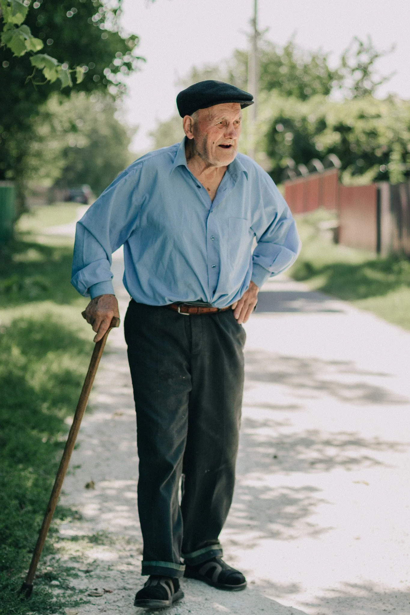 An elderly man with a cane standing on a sidewalk outdoors, wearing a light blue button-up shirt, dark pants, a black hat, and sandals, appearing to be looking at something with a surprised or curious expression.
