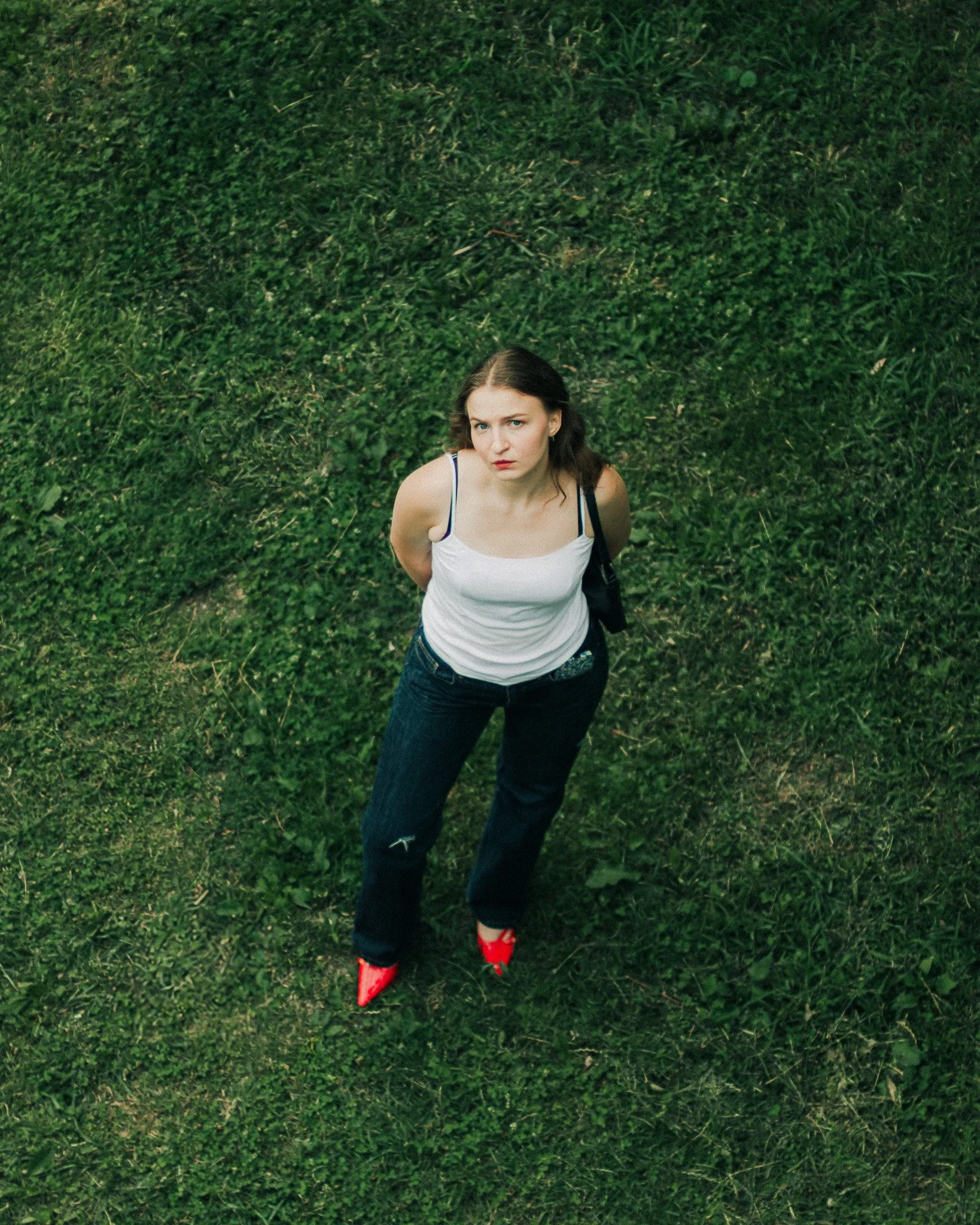 A young woman with brown hair, wearing a white tank top, dark jeans, and red high-heeled shoes, is standing on a grassy area, looking up at the camera with her hands behind her back.