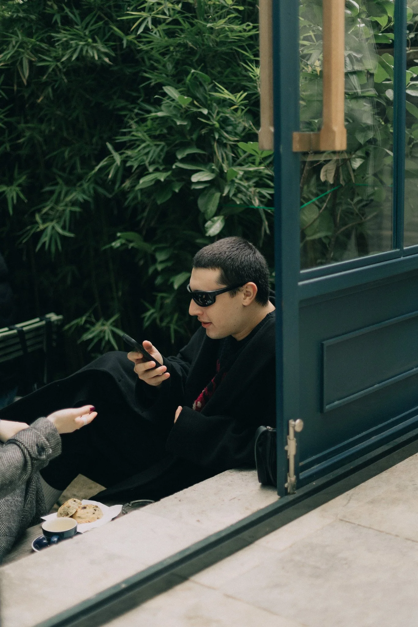 A man with short dark hair, wearing sunglasses and a black coat, sitting near a window with green foliage in the background, looking at his phone.