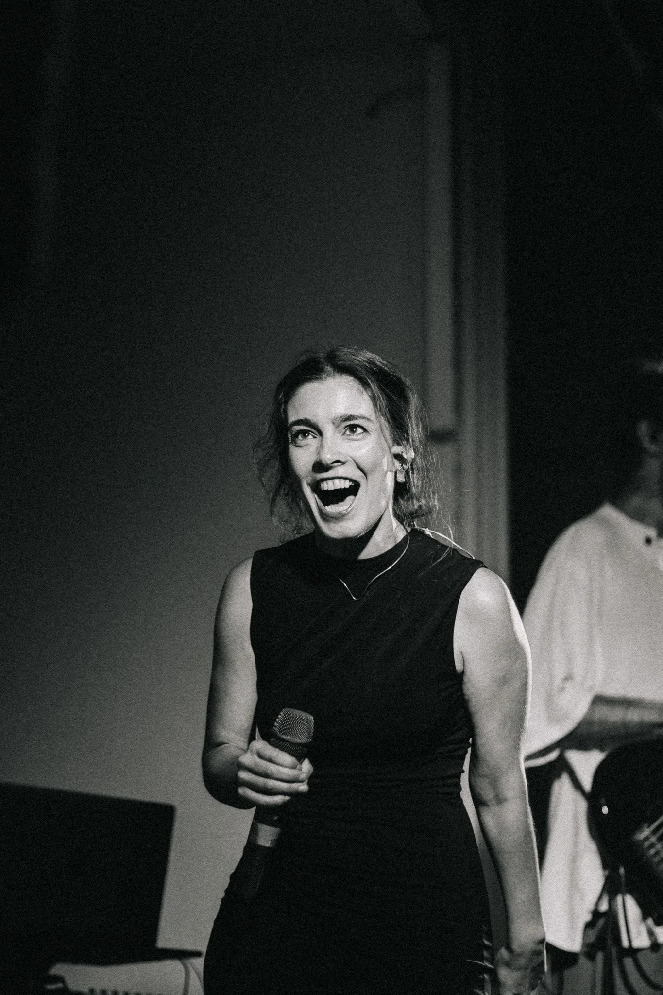A woman in a sleeveless dress holding a microphone, smiling and looking excited on stage. A person with glasses is in the background.