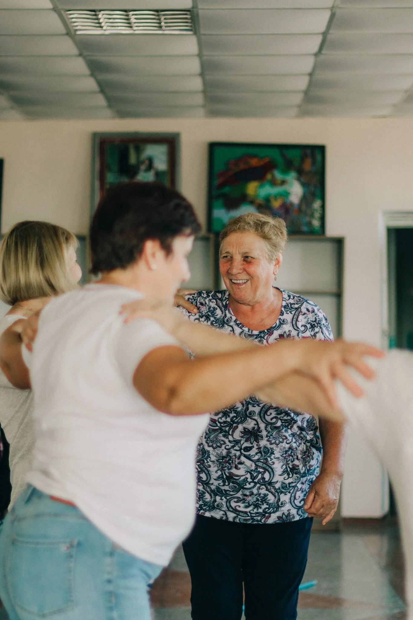 A group of people dancing and smiling indoors, including several women and a man, in a room with artwork on the walls.