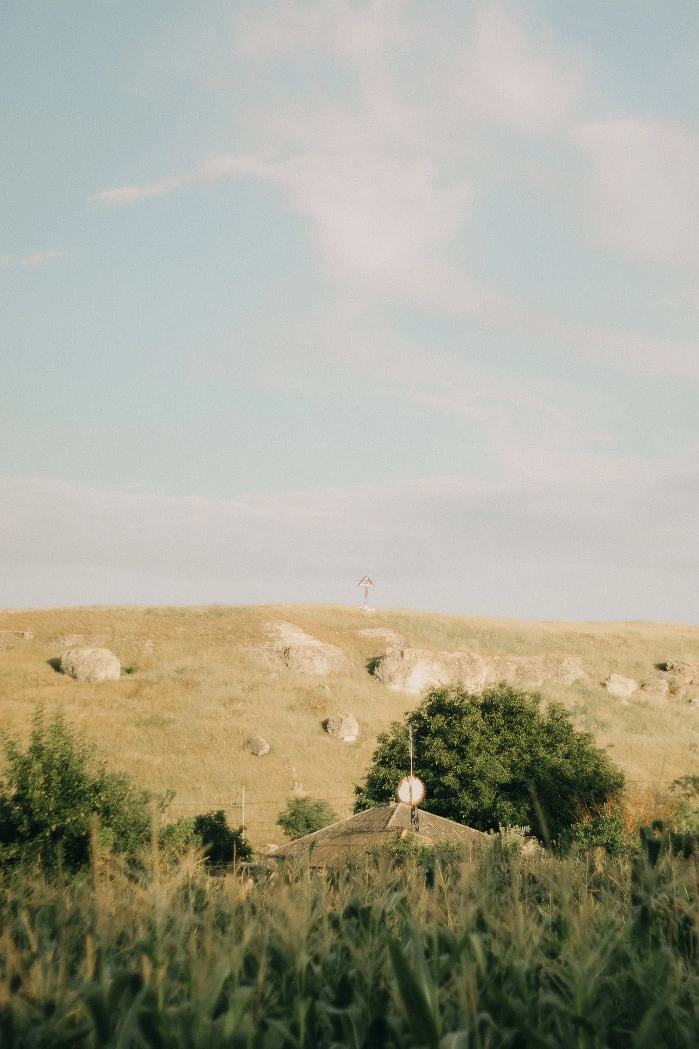 A rural landscape with a grassy hill, scattered rocks, a house with an antenna on its roof, and a large tree, under a partly cloudy sky.