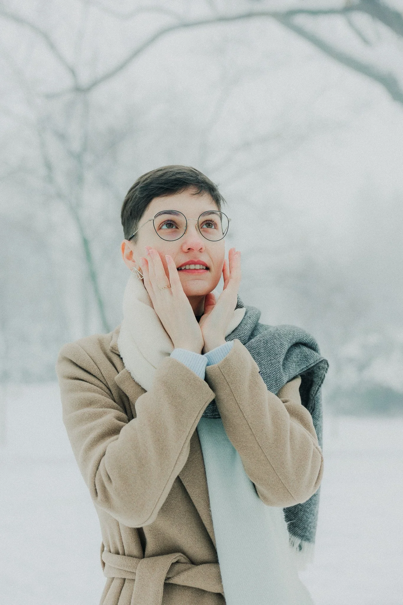 A woman with short dark hair and glasses in a scarf and coat standing outdoors in a snowy landscape, touching her face and looking up