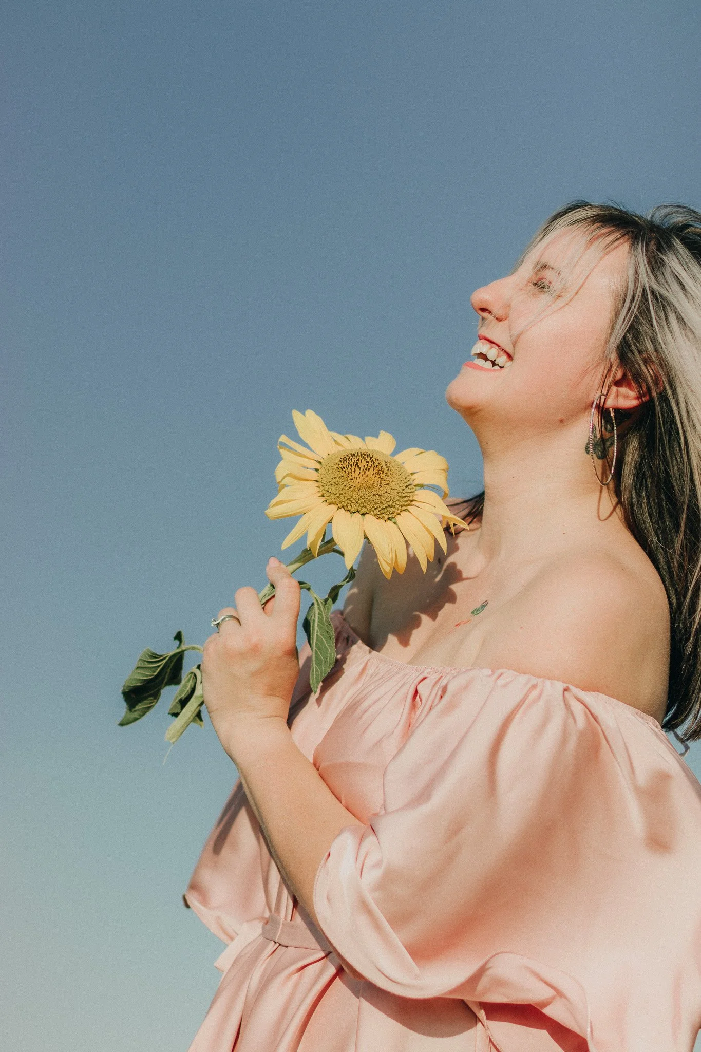 A woman with blonde hair holding a sunflower, smiling with her eyes closed, wearing a light pink off-shoulder dress and earrings, against a clear blue sky.