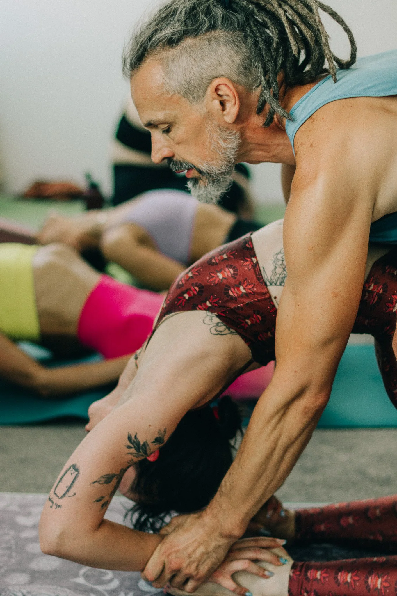 An older man leading a yoga class with a woman in wheel pose, focusing on her backbend in a yoga studio.