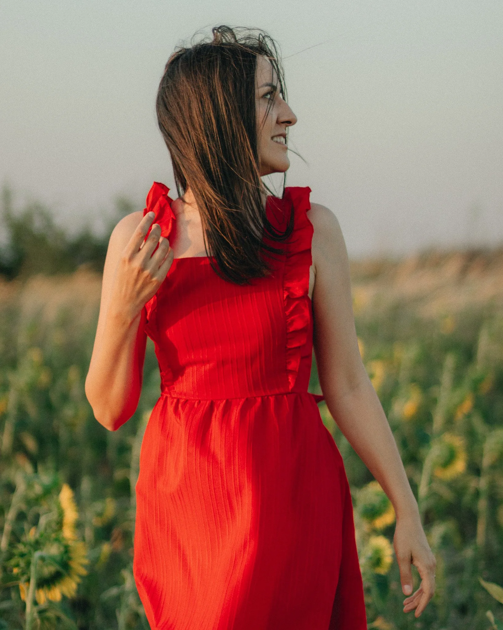 A woman in a red dress standing in a field of yellow flowers, looking to the side and smiling.