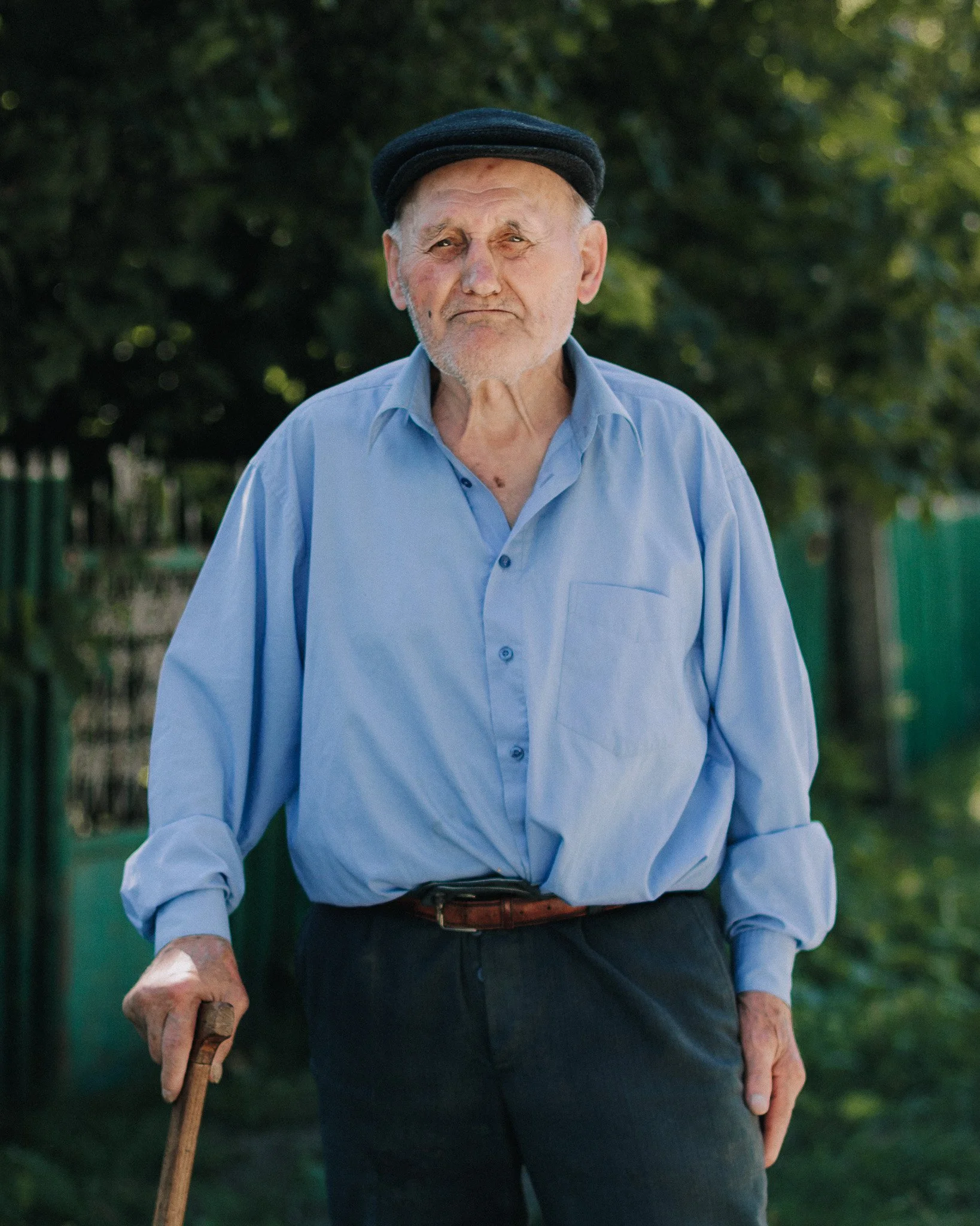 An elderly man with a white beard and mustache, wearing a black cap, light blue shirt, and dark pants, standing outdoors with trees in the background, holding a cane.