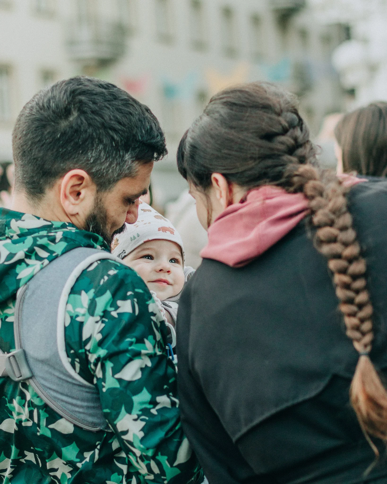 A family of three, consisting of a man, woman, and baby, sharing an affectionate moment outdoors on a city street, with buildings and blurry background visible.
