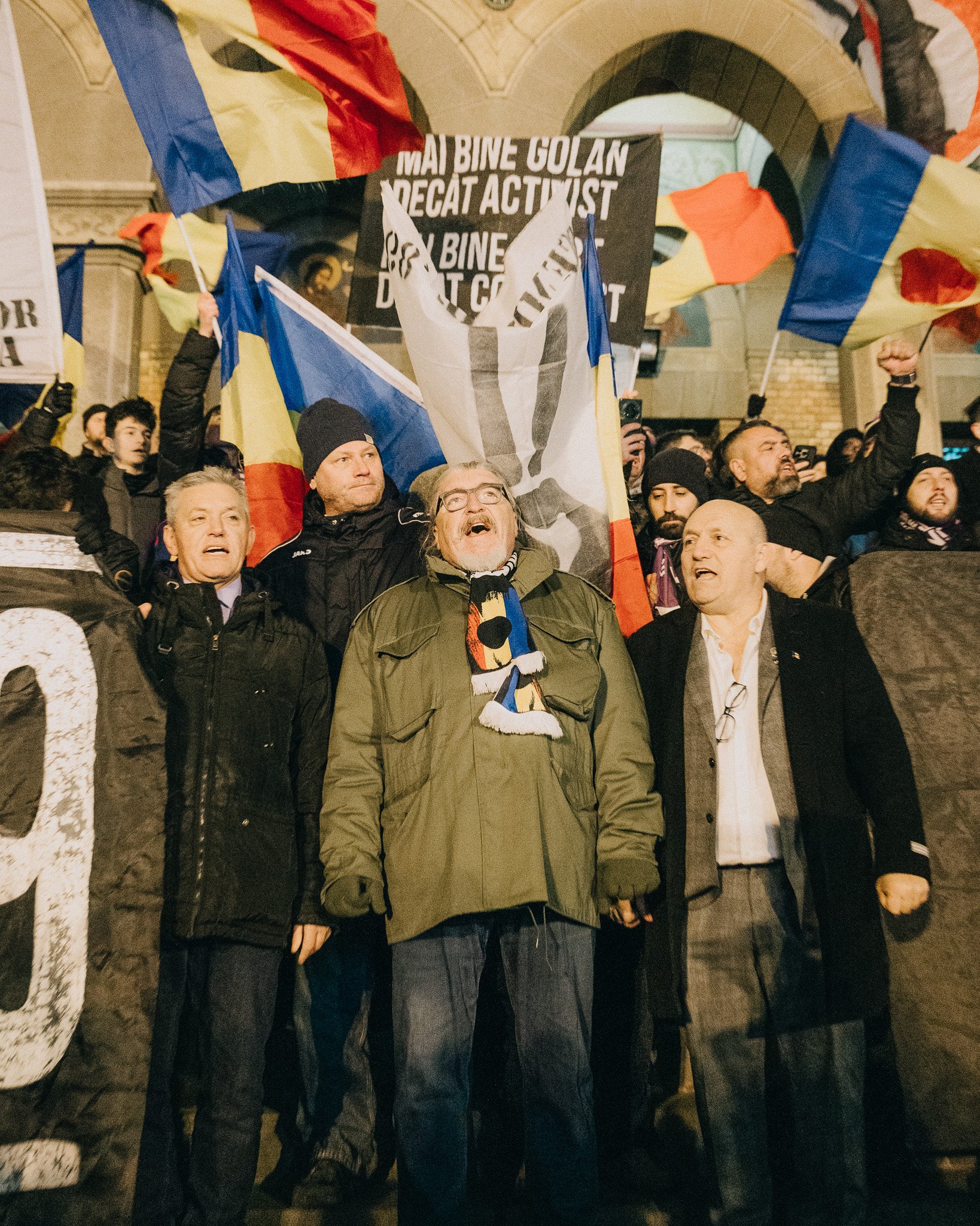 A group of protesters showing support with raised fists and flags, gathered outdoors at night, with a large banner in the background.