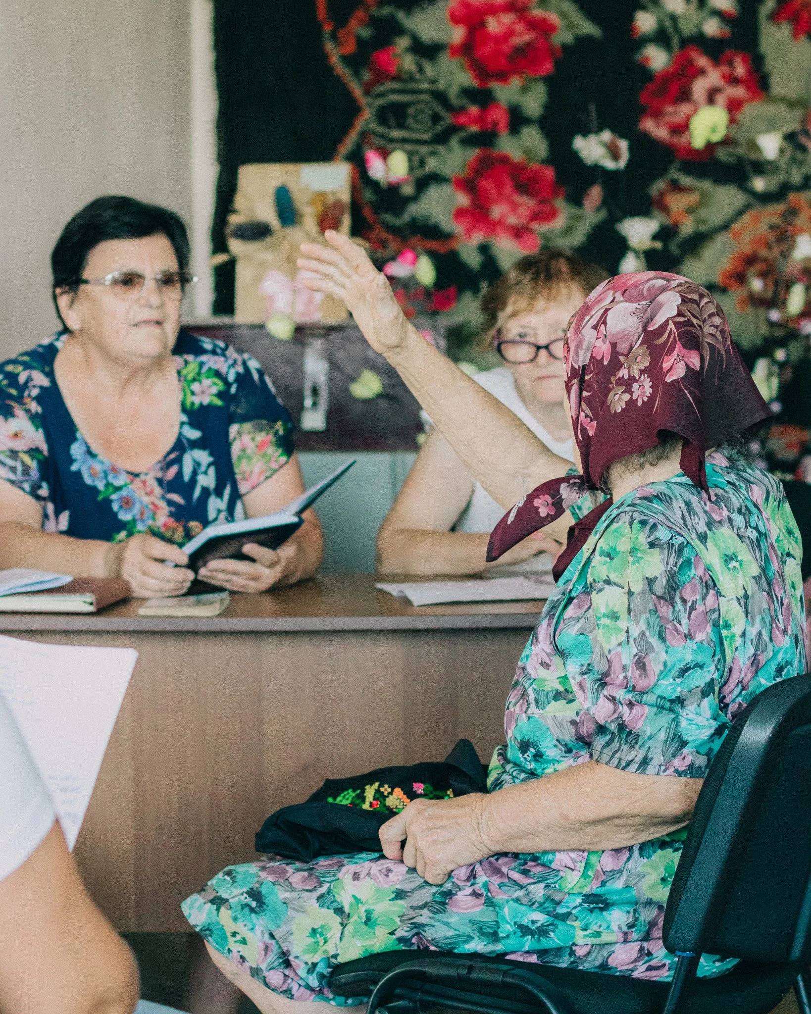 A woman wearing a floral dress and a headscarf sitting at a table raising her hand to participate in a discussion, with two other women listening attentively in a room decorated with a floral tapestry.