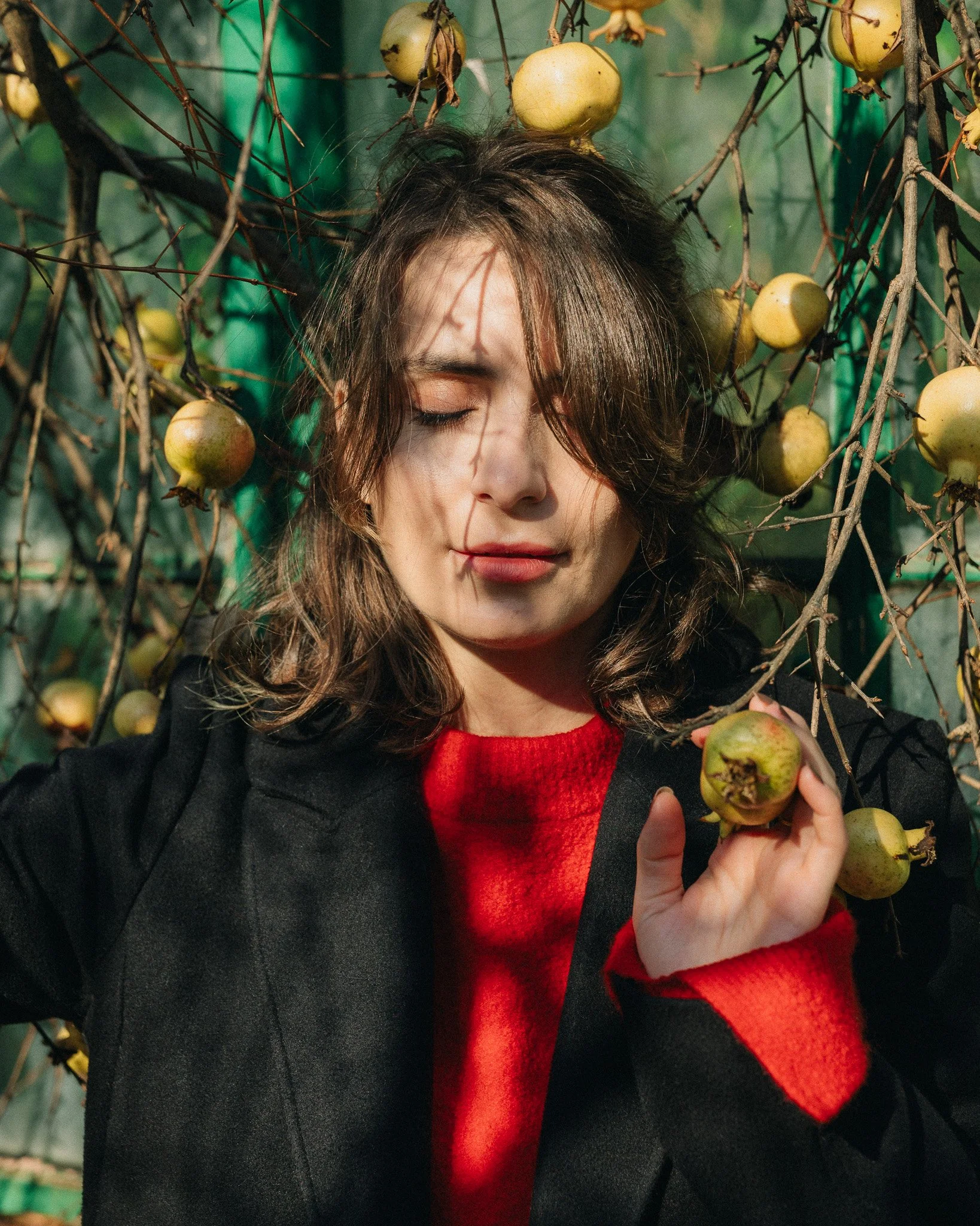 A young woman with wavy brown hair, wearing a red sweater and black coat, standing among bare trees with yellow apples hanging from the branches, with her eyes closed.
