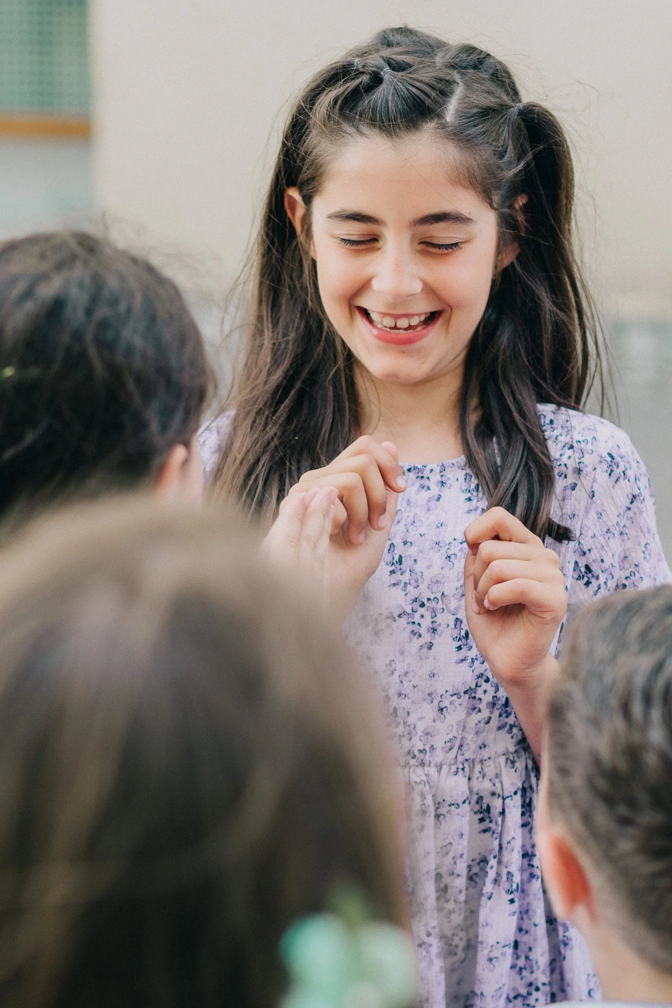 A young girl with long brown hair and a big smile, wearing a purple floral dress, is surrounded by other children in a classroom setting.