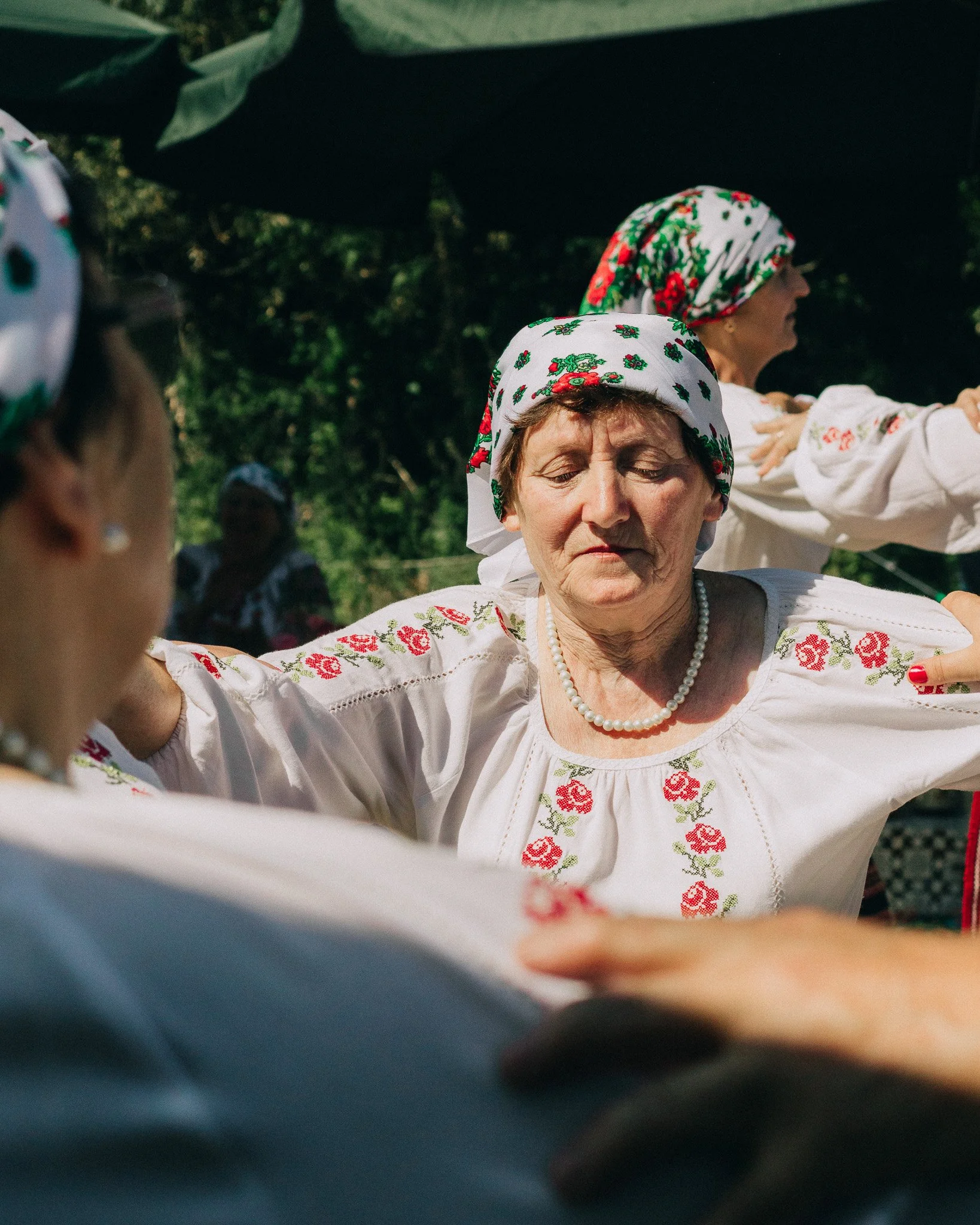 Older women in traditional embroidered blouses and floral headscarves participating in a dance outdoors.
