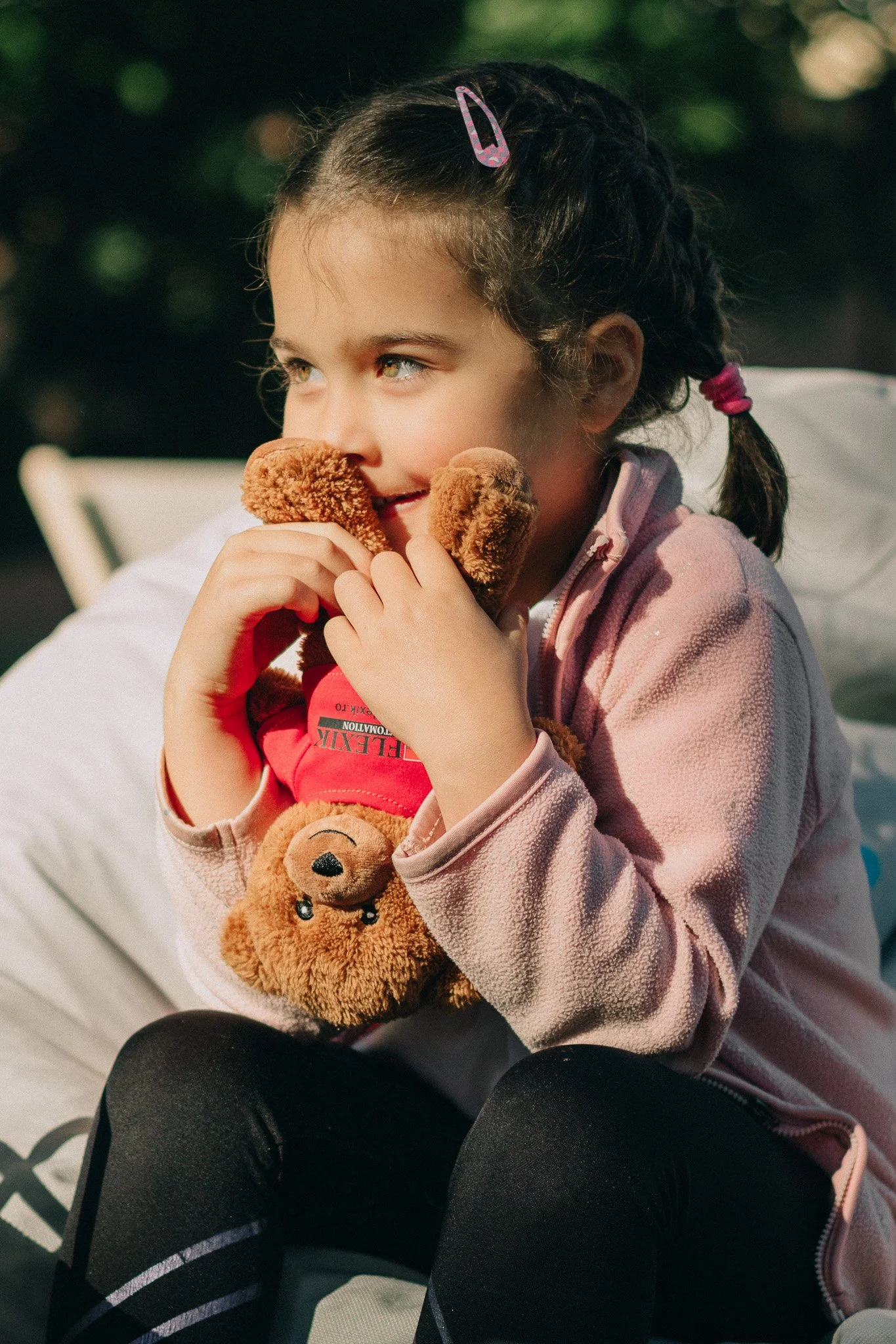 A young girl with dark hair in braids holding two teddy bears, sitting outdoors.