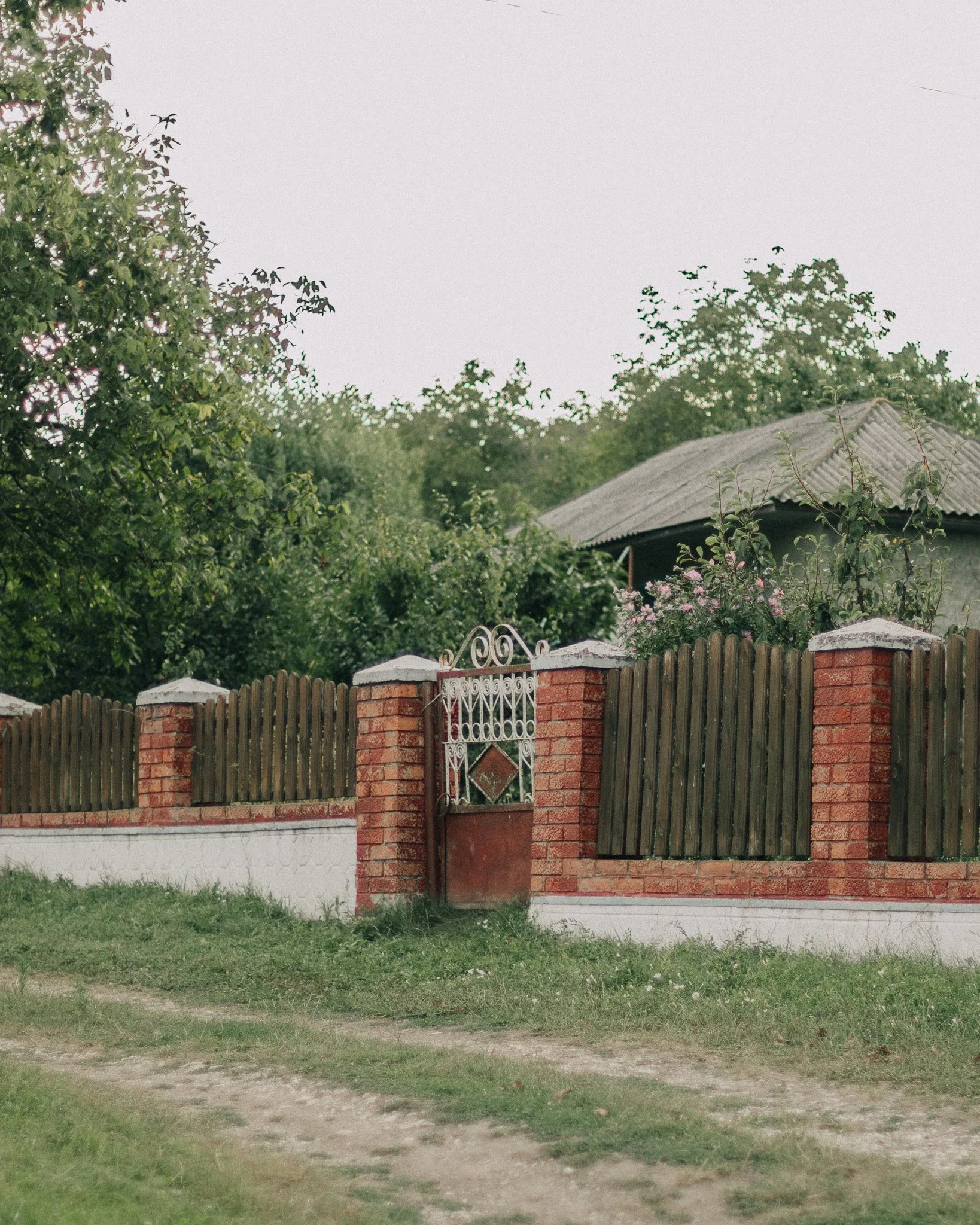 A brick and wooden fence with a metal gate surrounds a house with a gray roof, and trees and shrubs are visible behind the house.