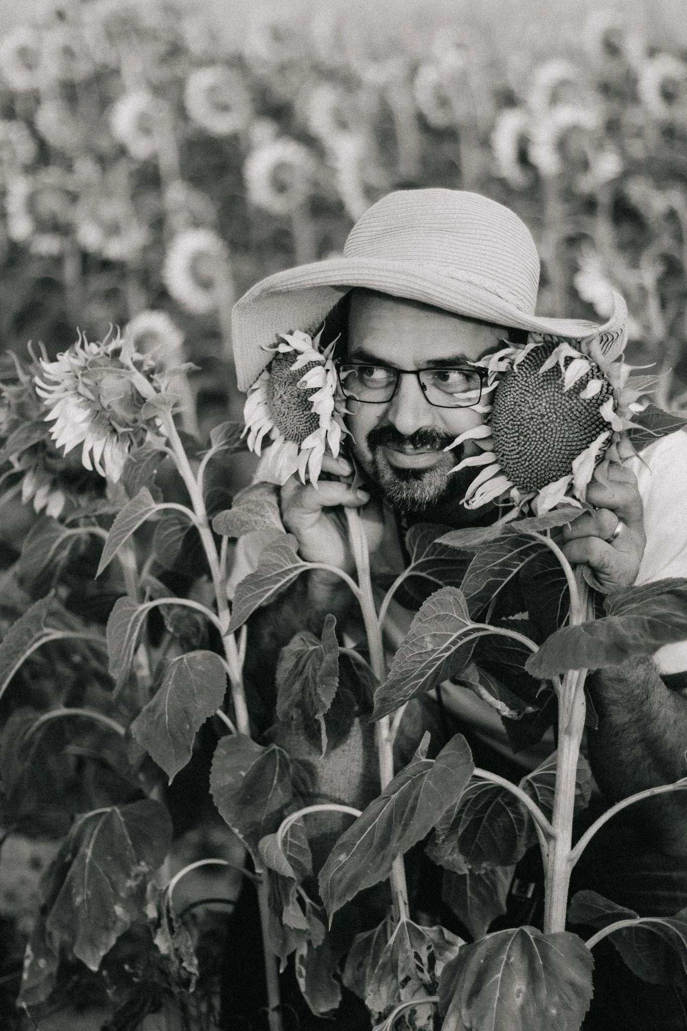 A man with glasses and a beard wearing a wide-brimmed hat, holding sunflower heads near his face, surrounded by sunflower plants in a field.