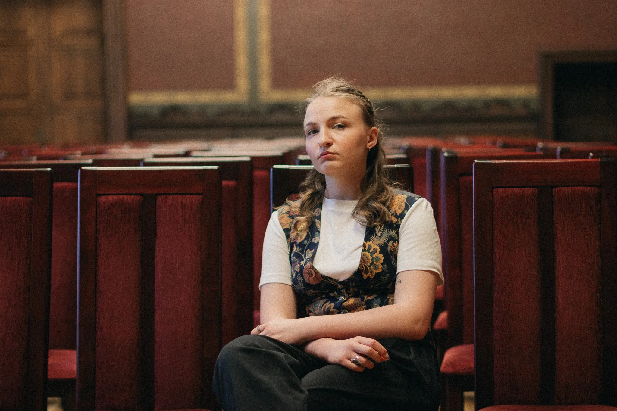 Young woman sitting alone in an empty theater or auditorium with rows of red cushioned seats, wooden paneling, and a decorative wall in the background.