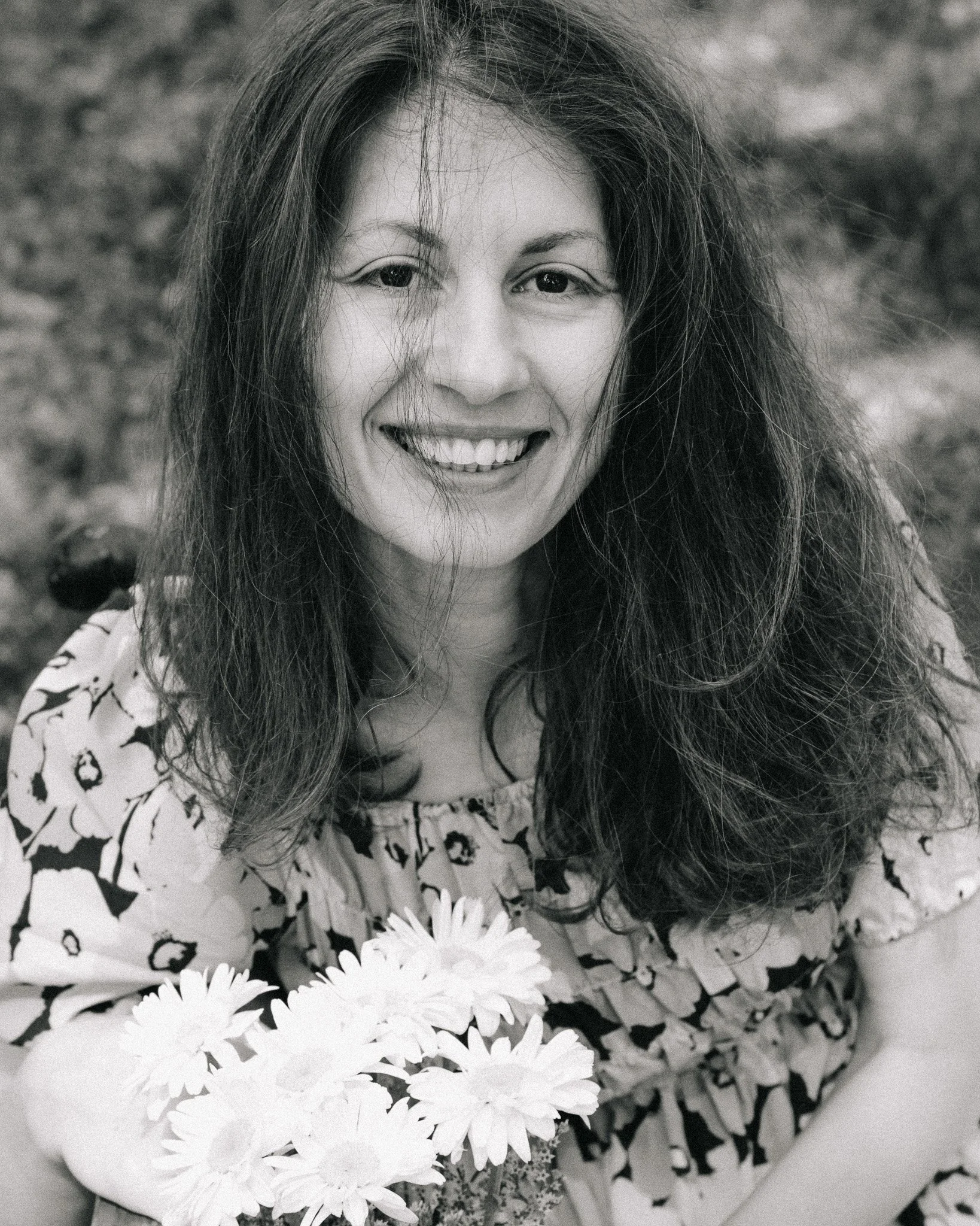 A woman with long hair smiling while holding a bouquet of flowers, wearing a floral dress.
