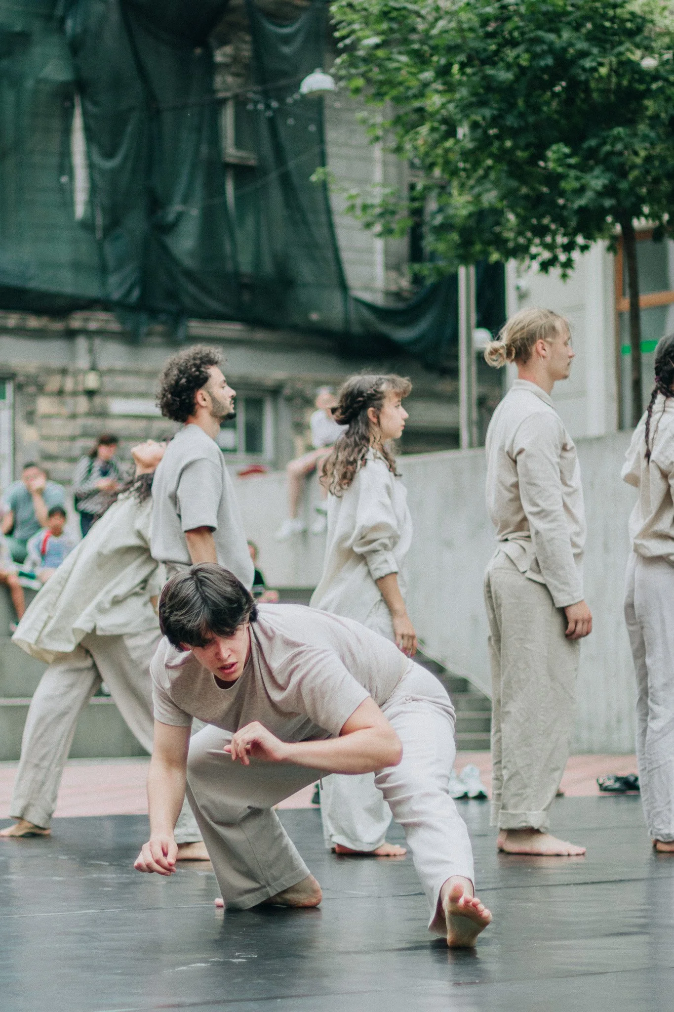 A group of people practicing martial arts or dance outdoors, with some in a kneeling or standing position, on a black mat, surrounded by onlookers and greenery.