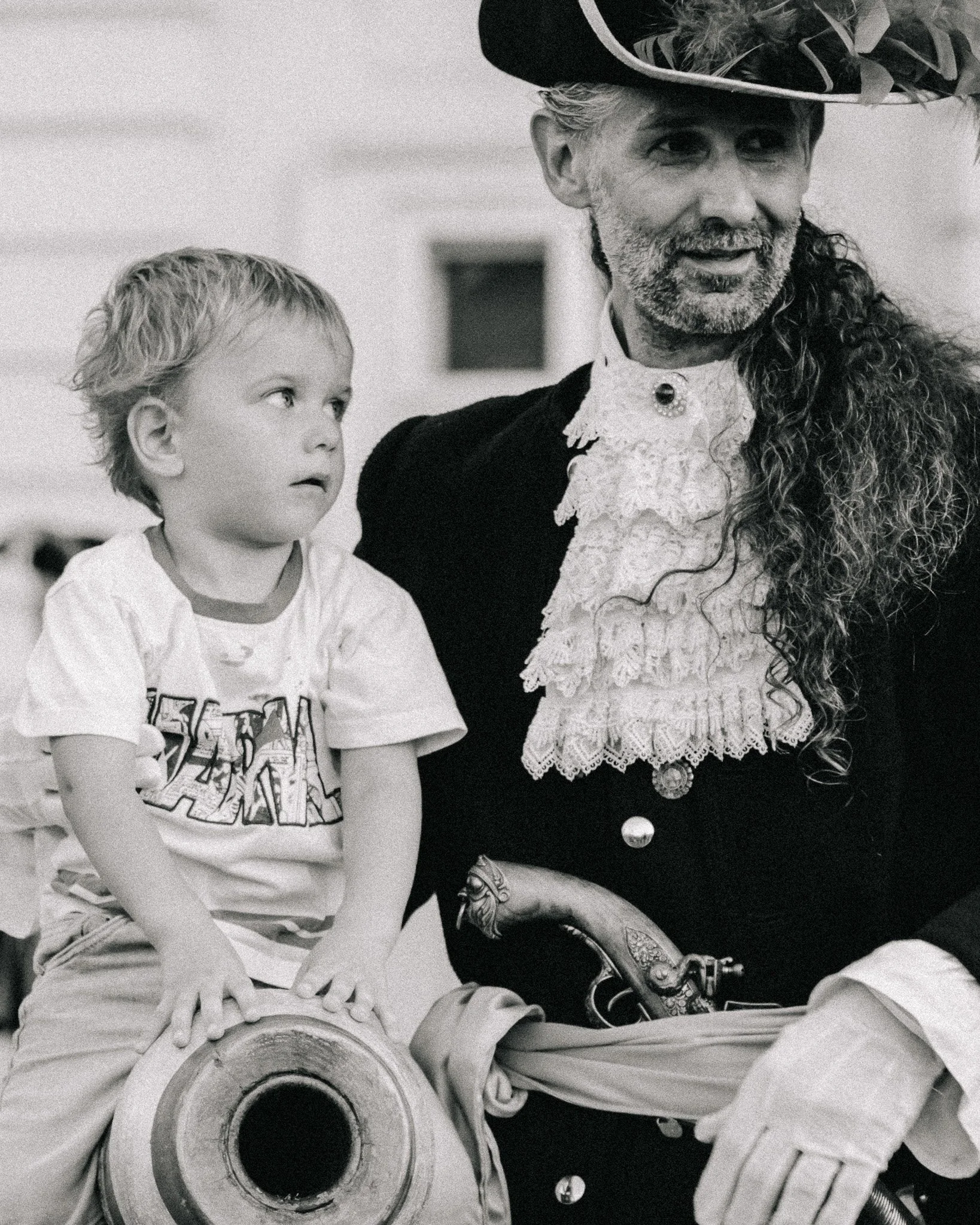 A young boy sitting on a decorated cannon, looking to the side, with a man dressed as a historical figure, possibly a pirate, holding the cannon. The man has a long curly beard, a wide-brimmed hat, and elaborate clothing with lace.