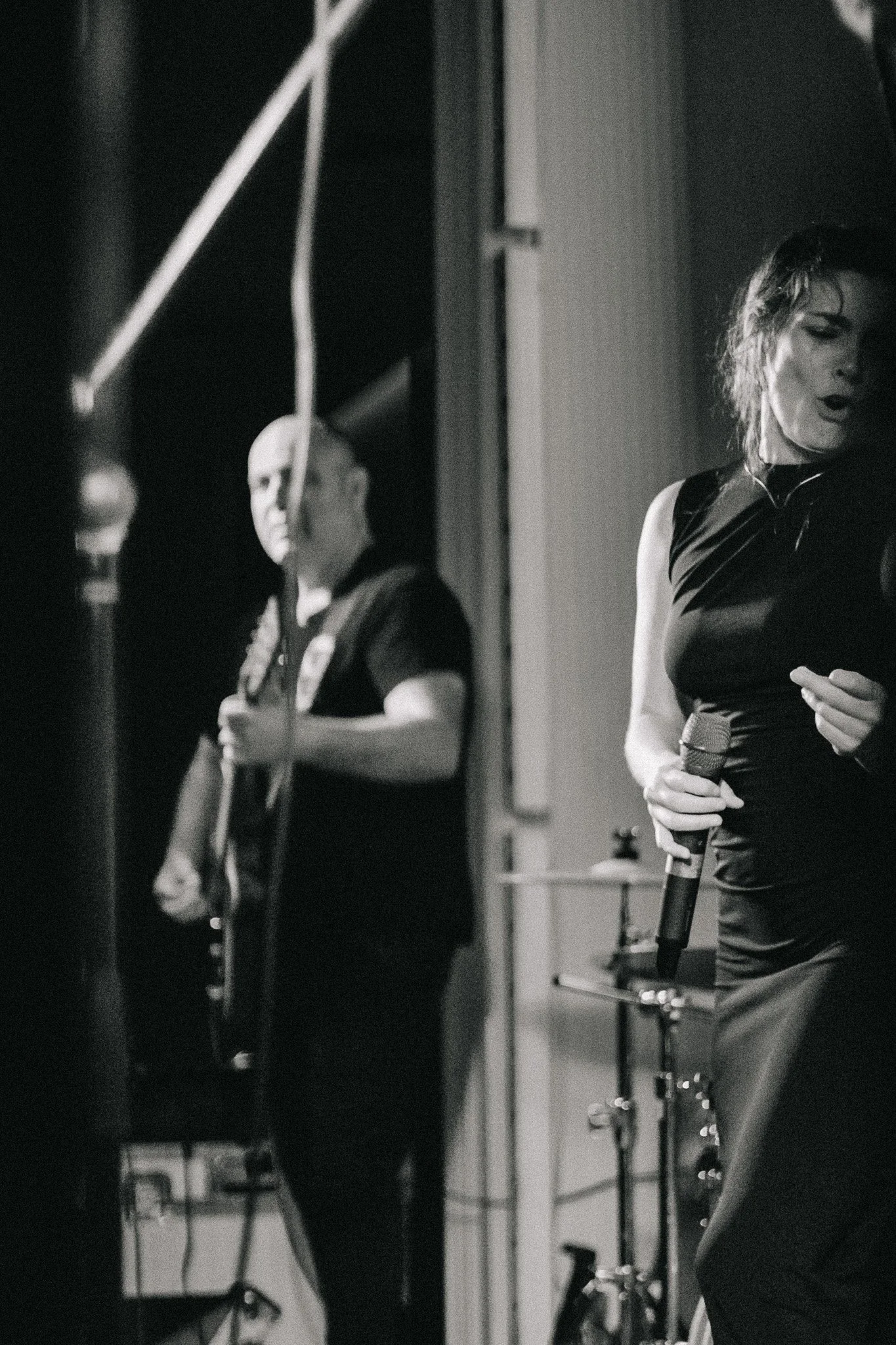 Black and white photo of a woman singing into a microphone on stage, with a band member playing guitar in the background.