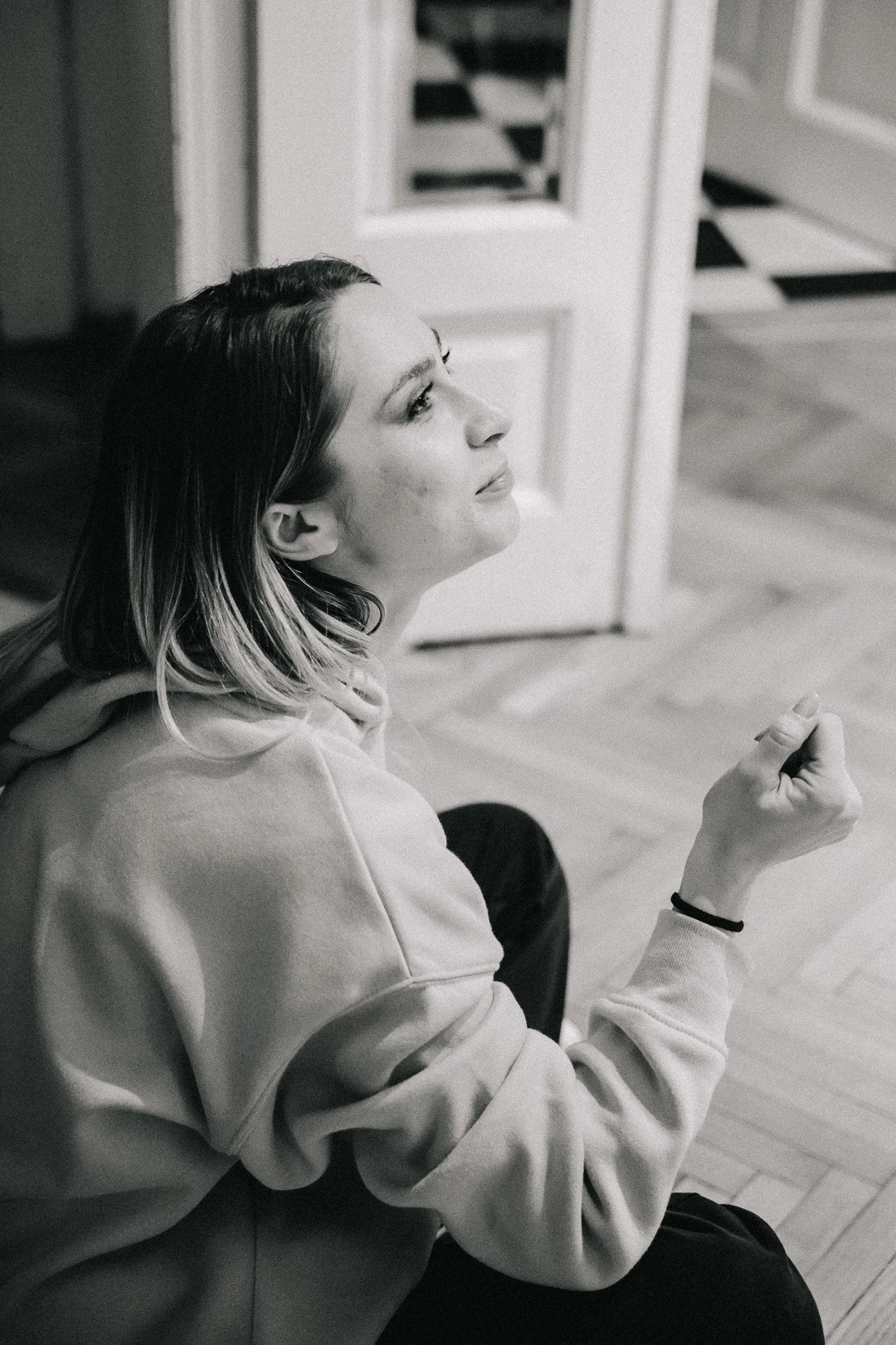 A black and white photo of a woman with shoulder-length hair, sitting on the floor in a room with wooden flooring, wearing a hoodie, making a fist with her right hand, looking contemplative.