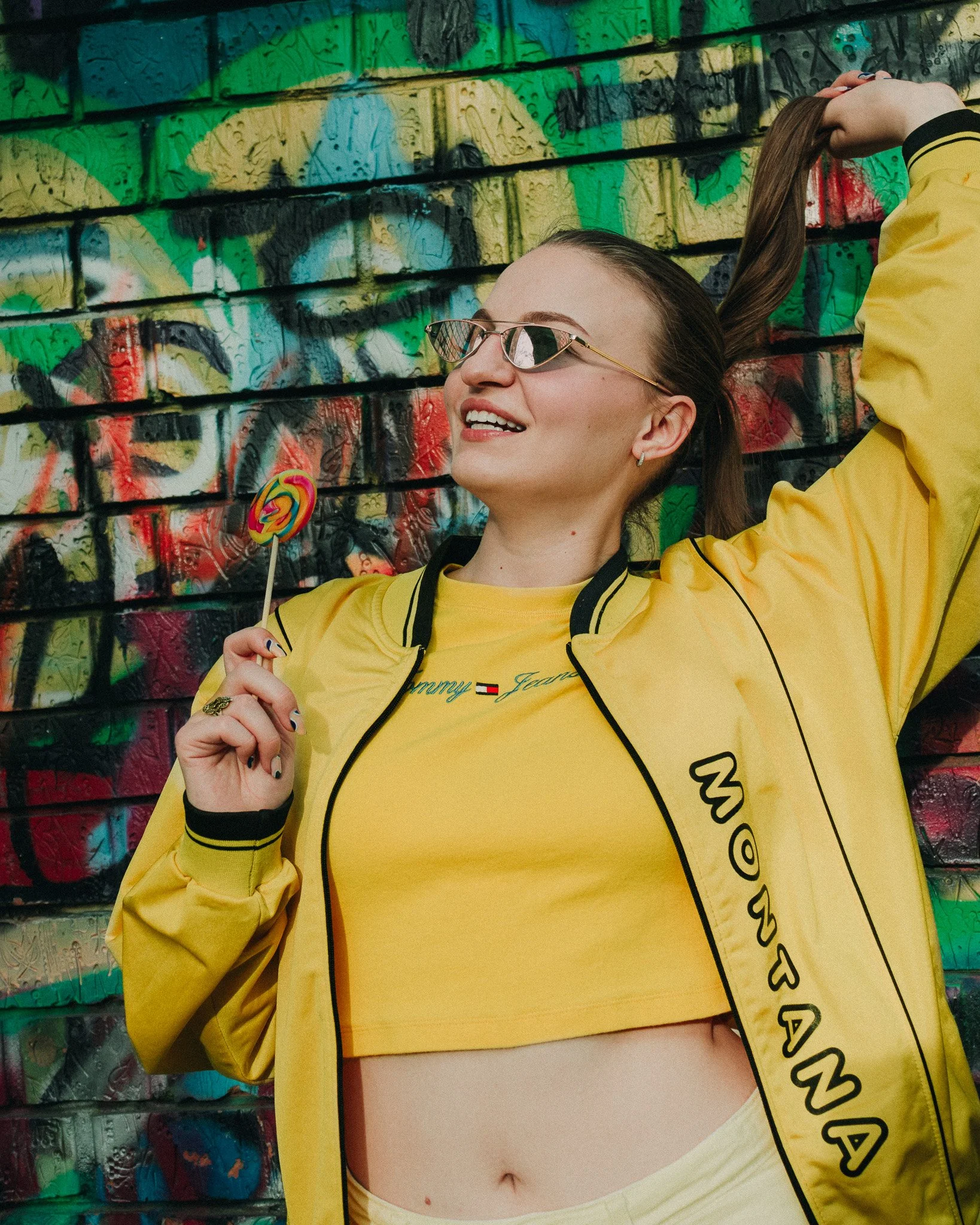 A woman wearing yellow Tommy Hilfiger athletic wear and sunglasses, smiling while holding a colorful lollipop, standing against a graffiti-covered wall.