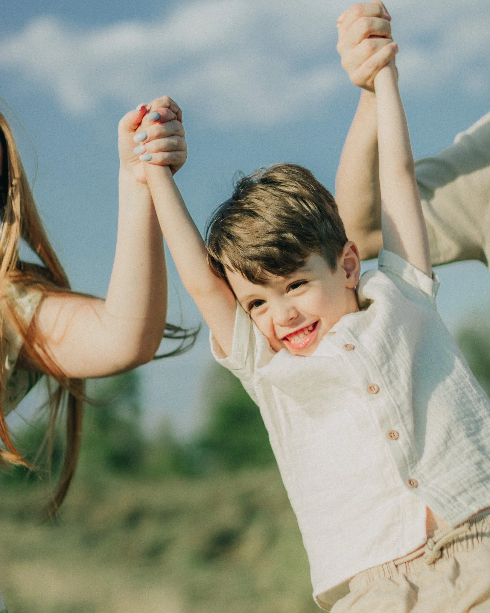 A smiling young boy being lifted in the air by adults, holding their hands, outdoors on a sunny day.