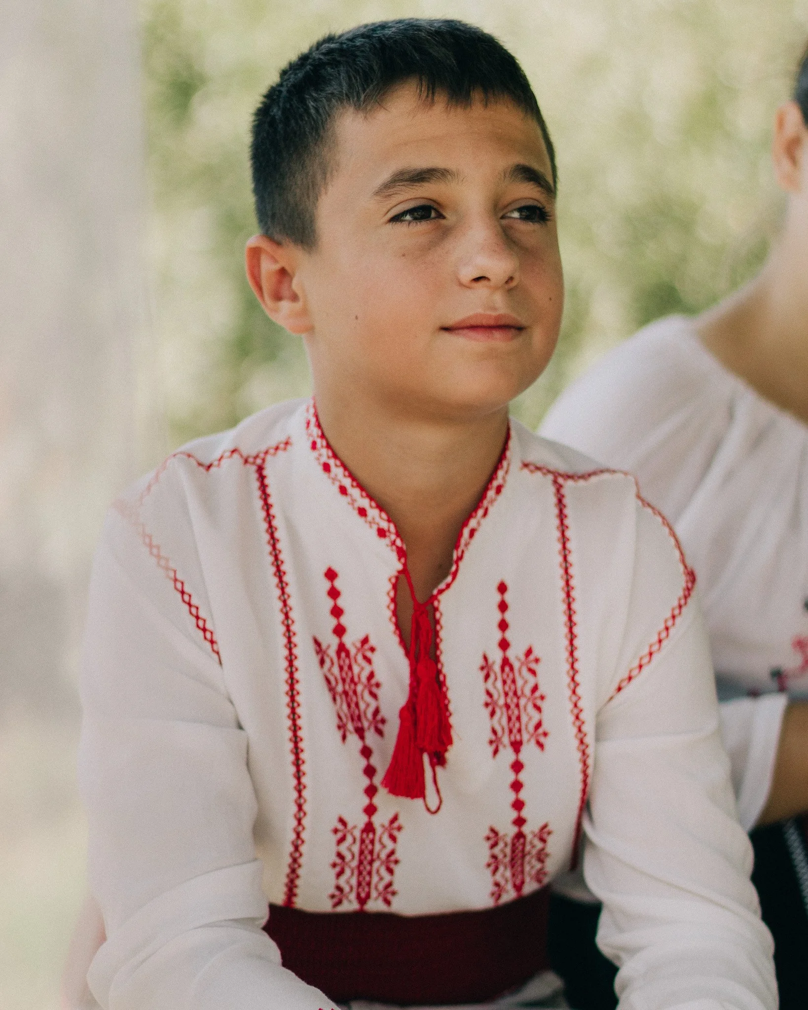 A boy wearing a traditional embroidered white shirt with red patterns and tassel, sitting outdoors.