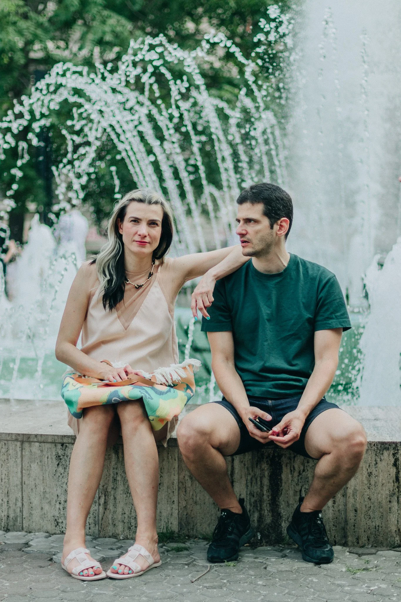 A woman and a man sitting on the edge of a fountain in a park, with water jets behind them. The woman has a colorful bag on her lap and is wearing a sleeveless top and sandals. The man is wearing a dark green t-shirt, shorts, and sneakers, holding a 