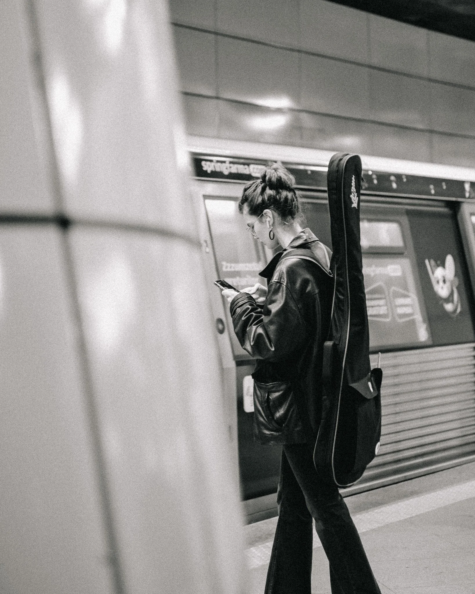 A woman with glasses and earrings, dressed in a leather jacket and pants, is standing on a subway platform looking at her phone, with a guitar case strapped to her back.