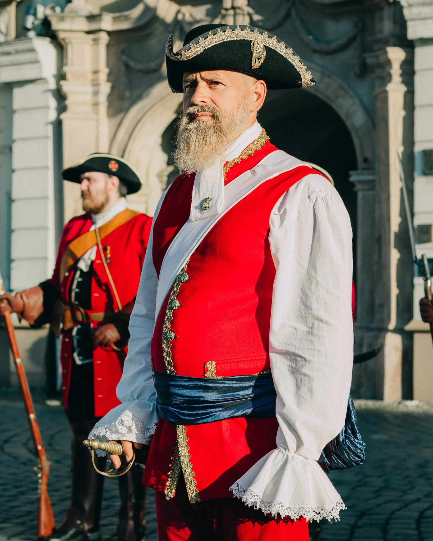 Two men dressed in historical military uniforms, with the man in the foreground wearing a red coat with gold embroidery, white lace cuffs, and a black tricorn hat with silver trim. The background features a similar figure and a stone building.