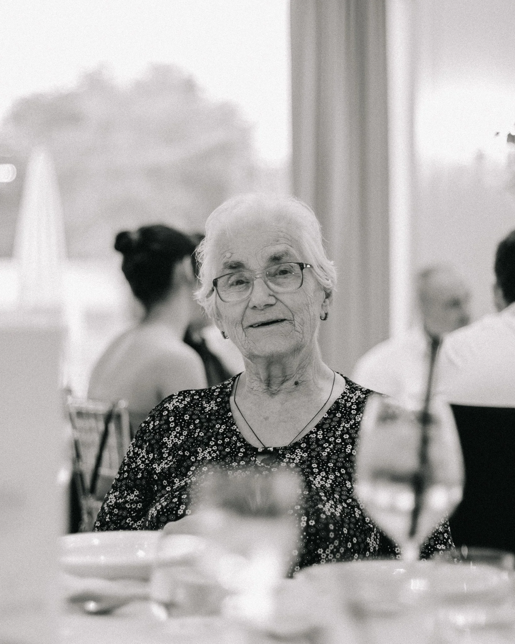 An elderly woman with glasses and earrings, smiling at a dining table during a gathering or celebration.