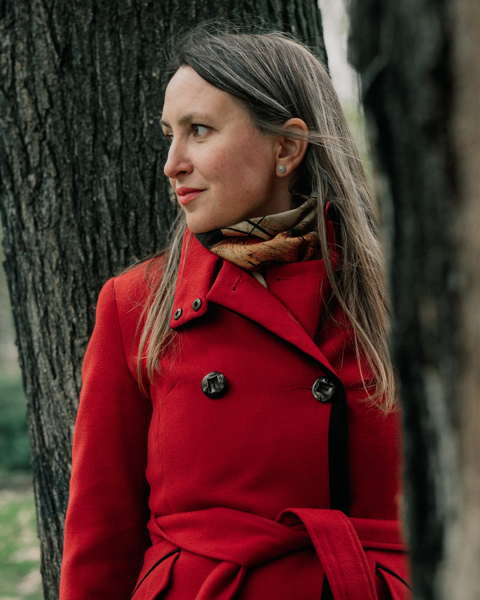 A woman with long brown hair, wearing a red coat and scarf, standing outdoors next to a tree, looking to the side.