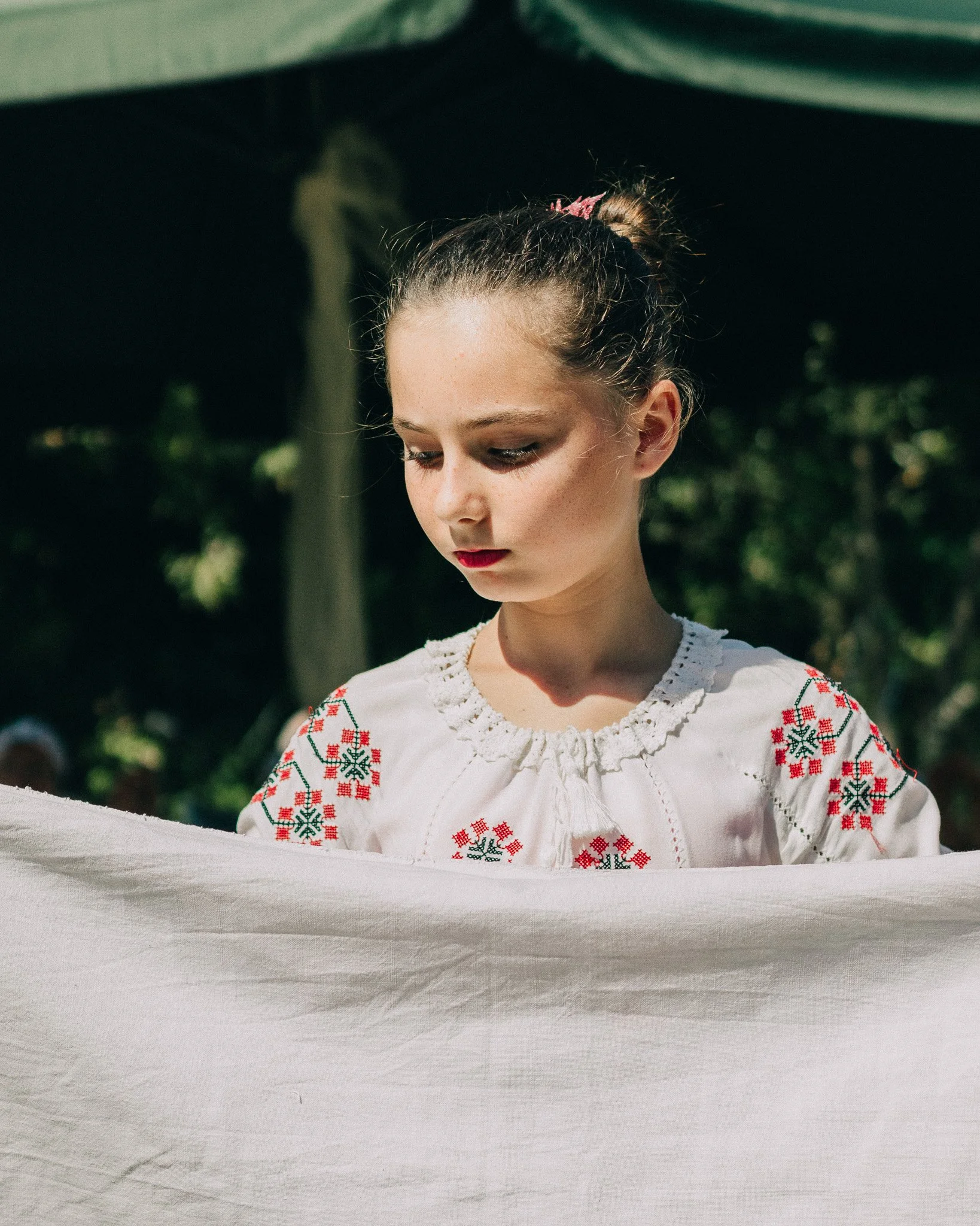 Young girl wearing traditional embroidered blouse, standing outdoors, looking down, with a white fabric in front of her.